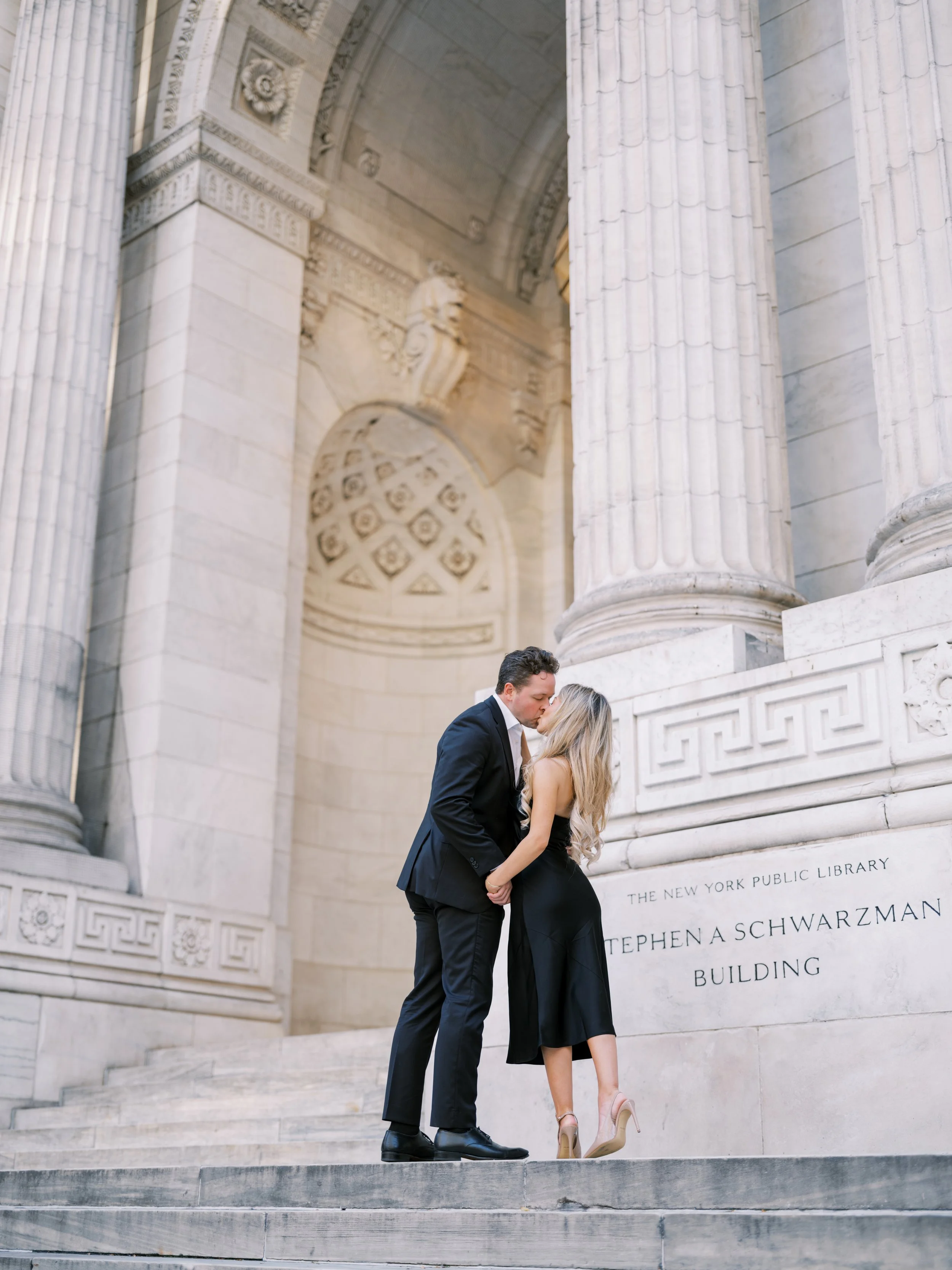 A man and woman kissing on stone steps in front of a historic building with large columns and detailed architectural features.