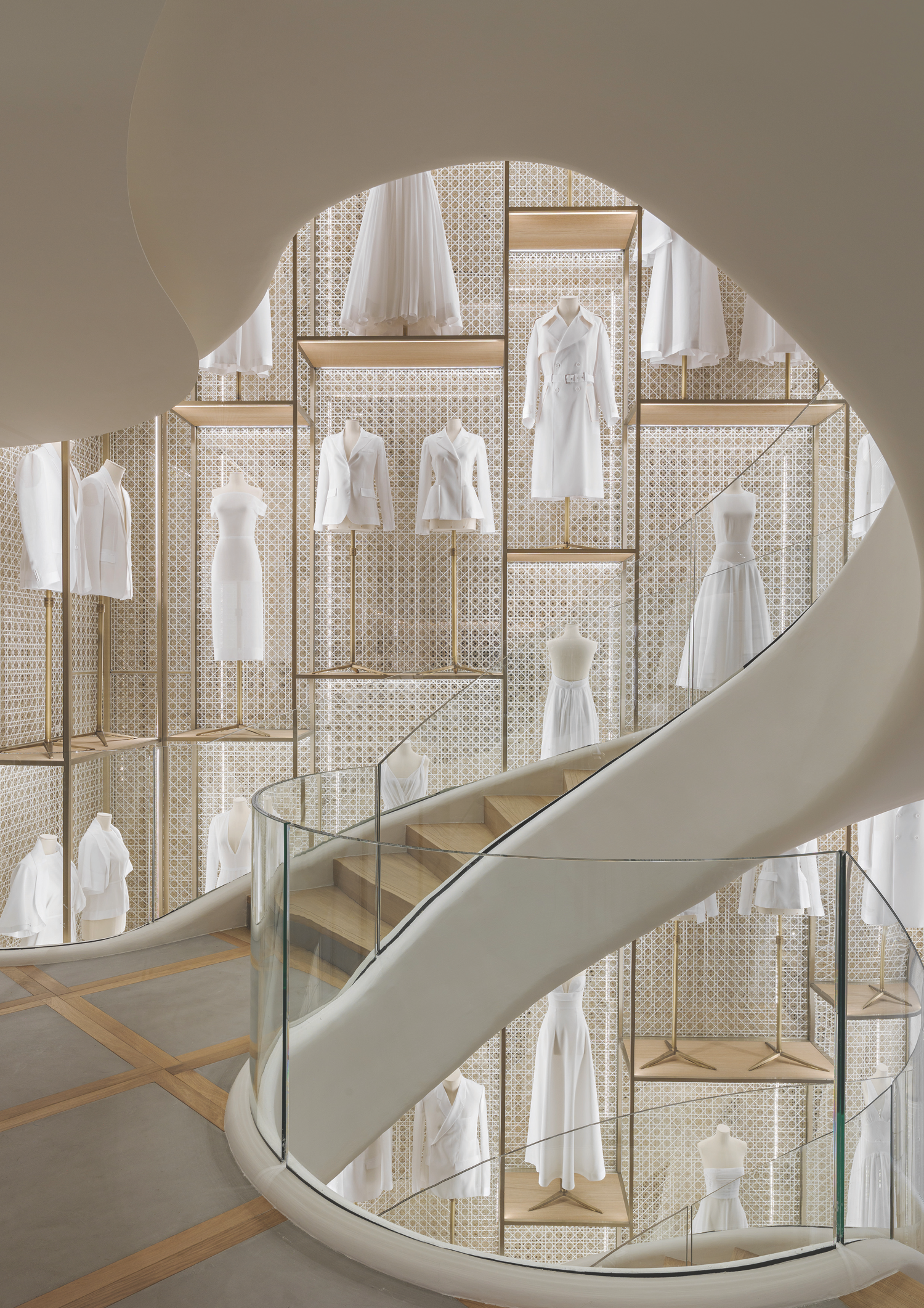 Display of white dresses, overcoats, and blouses on mannequins and clothing racks arranged on a decorative lattice wall in a modern boutique or showroom, viewed through a spiral staircase with glass railing.