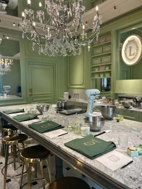 Luxurious kitchen and dining area with a large marble island, green cabinetry, and a crystal chandelier overhead. Items on the island include a stand mixer, bowls, and napkins, with seating around the counter.