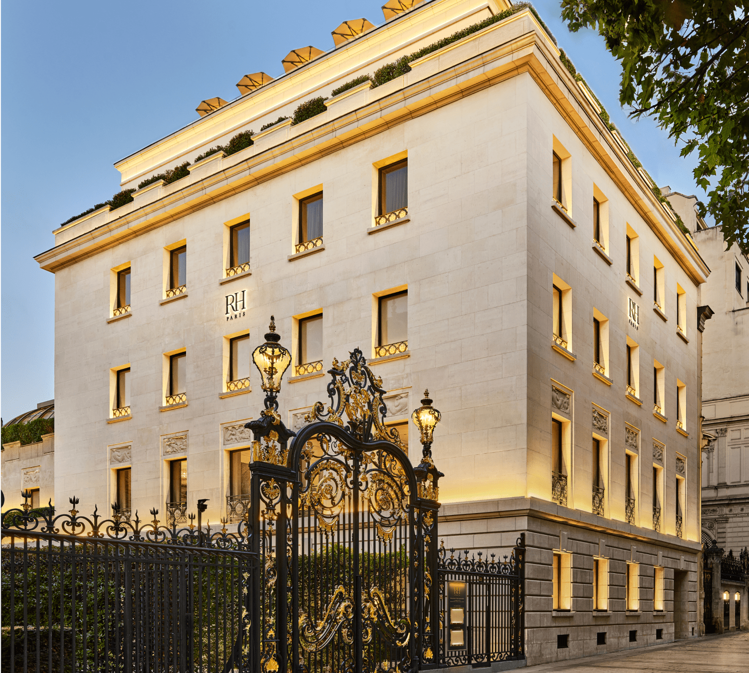 A multi-story luxury hotel building with the sign 'RH Paris' on the facade, surrounded by ornate wrought iron gates and street lamps, with trees and a clear blue sky in the background.