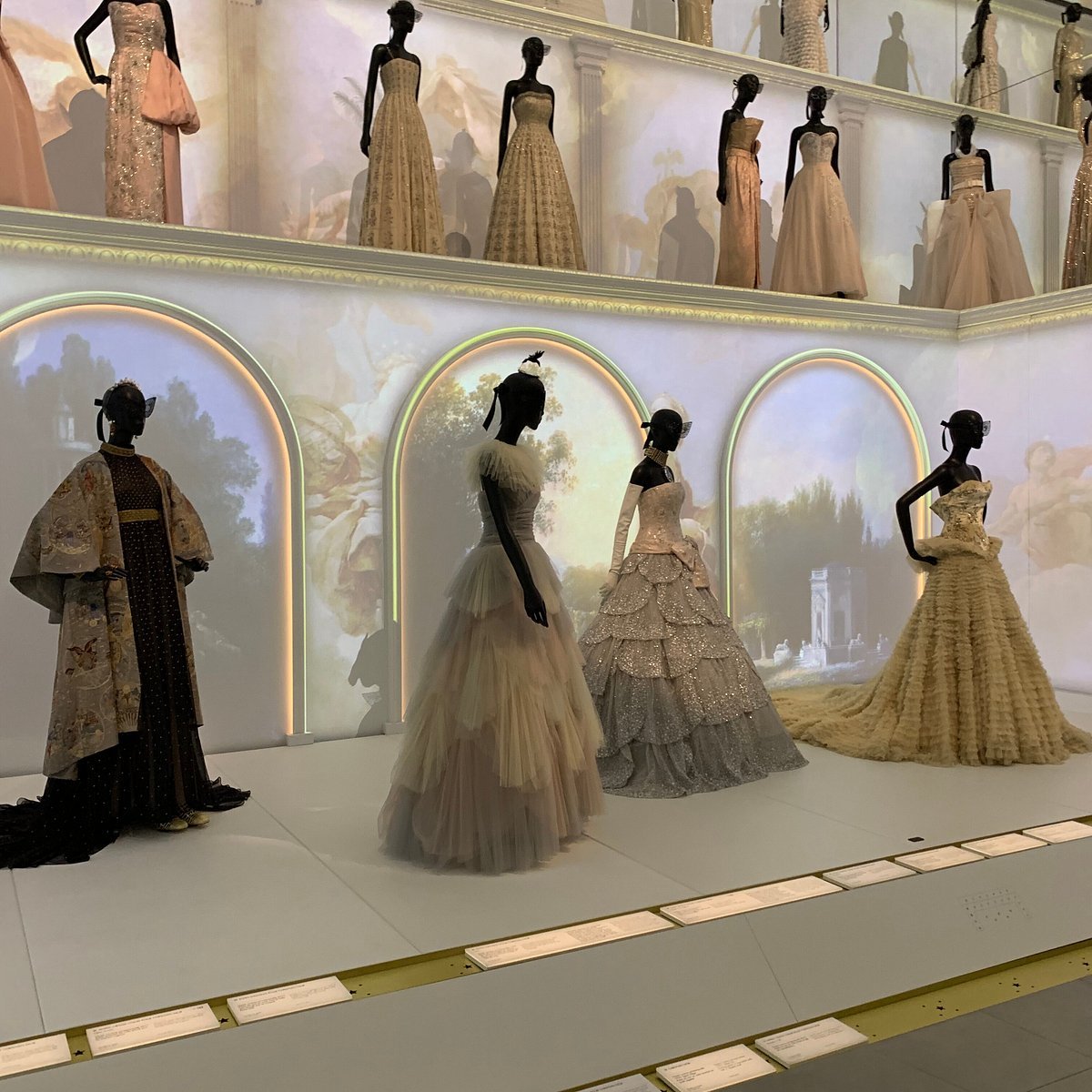 Display of mannequins dressed in elaborate, historical-style evening gowns on two levels, with soft background lighting and scenic backdrop images.