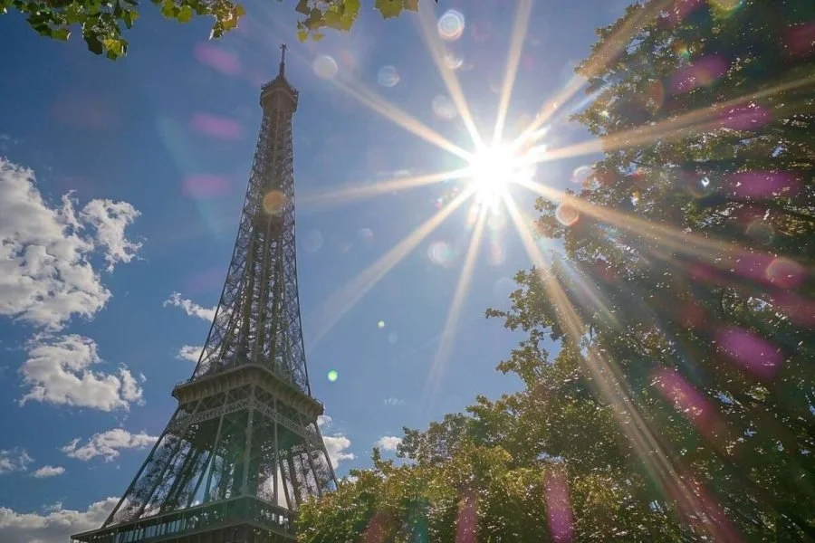 The Eiffel Tower with a bright sun in the sky and trees nearby.
