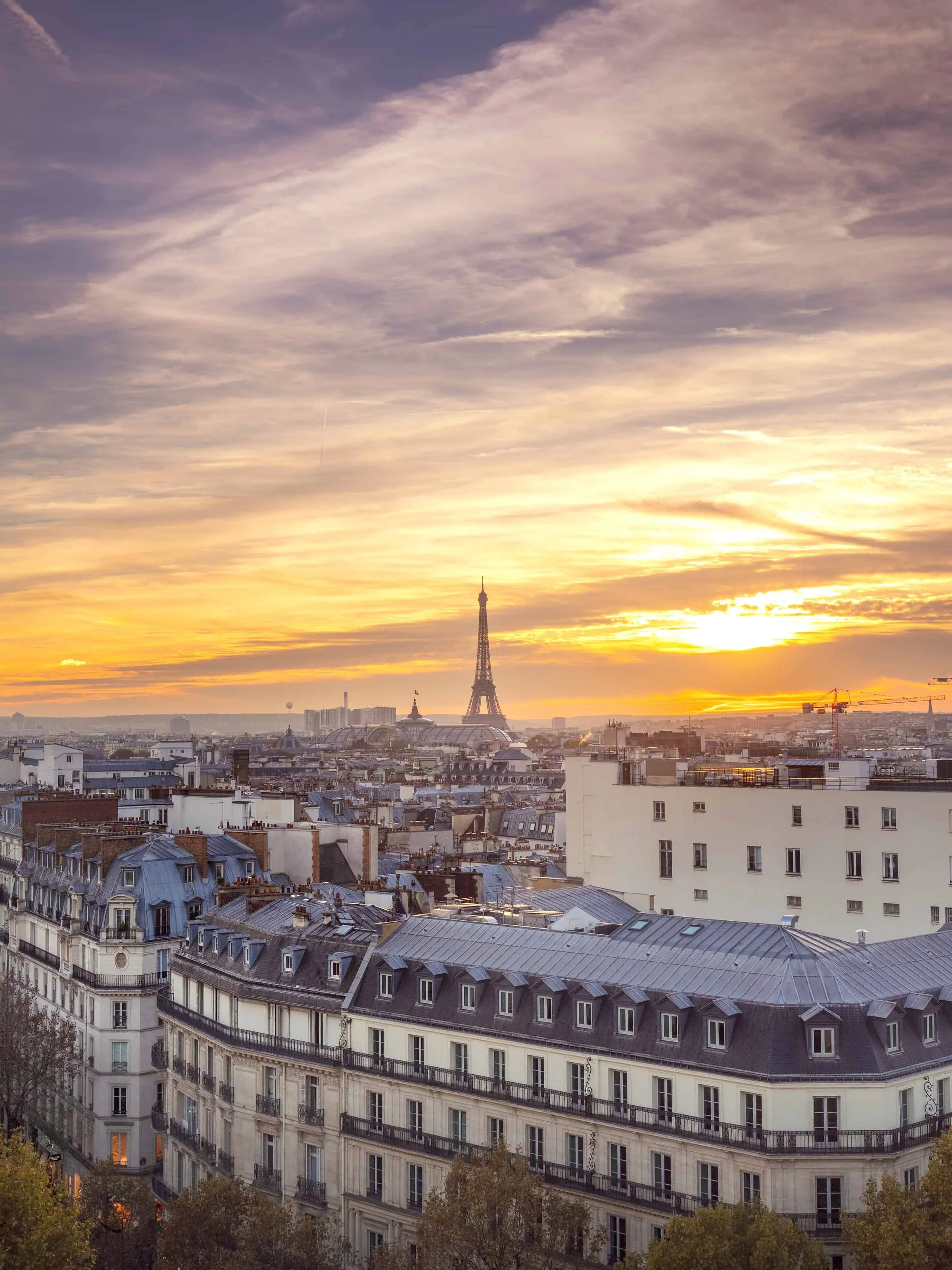 Cityscape of Paris with the Eiffel Tower at sunset. Rooftops and buildings in the foreground, sky with clouds and warm light.