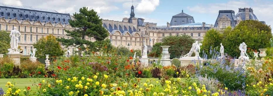 Ornate garden with statues, colorful flowers, neatly trimmed lawns, and historic buildings in the background.