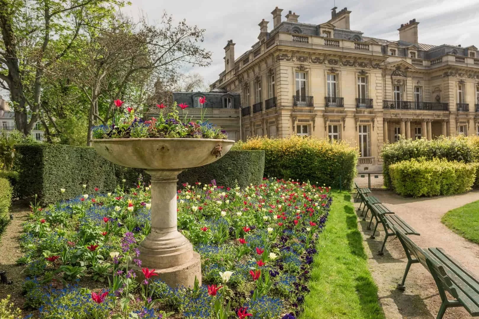 A historic building behind a flower garden with a large stone planter, benches, trees, and a pathway.