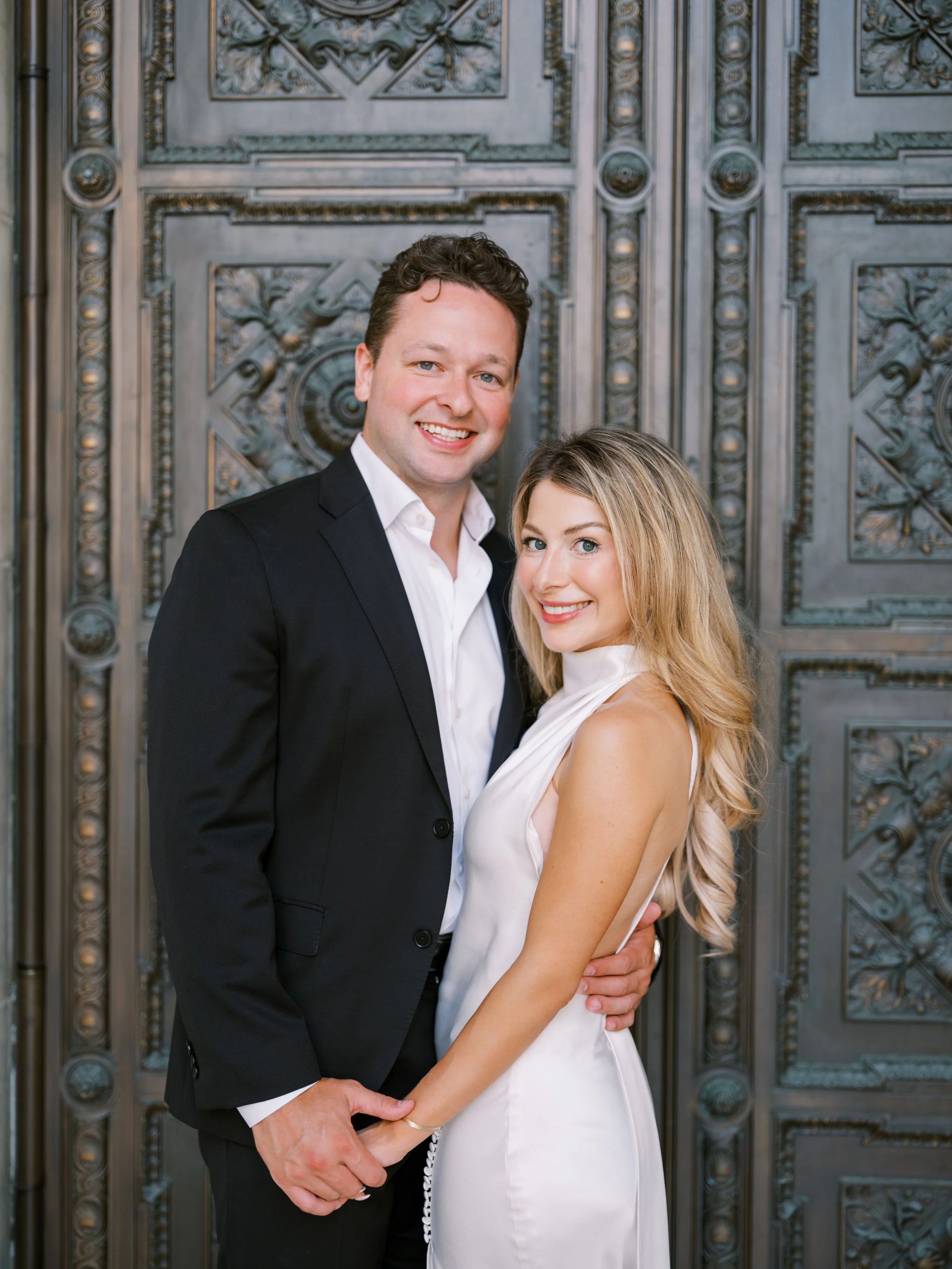 A smiling young man and woman in formal attire, holding hands in front of an ornate metal door with intricate designs. New York Public Library. NYPL. Samantha Daniell & Michael Moore. 