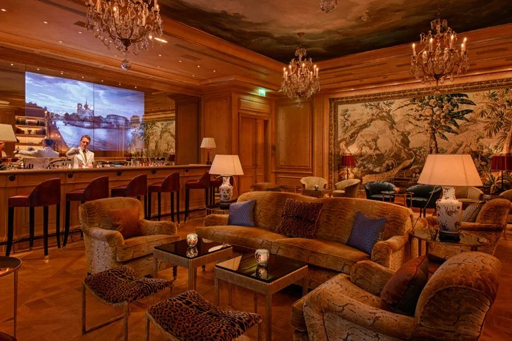 Luxurious lounge with wooden paneling, vintage furniture, chandeliers, and a large tapestry wall hanging. A bar with chairs is on the left, and a large LED screen displays a river scene.