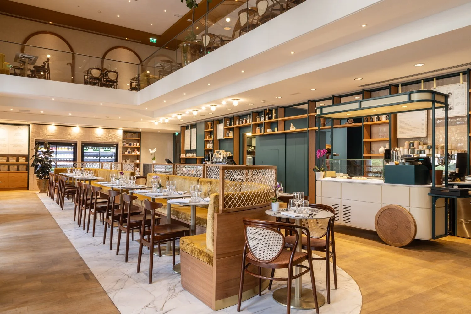 Elegant restaurant interior with a long dining table set with glasses and silverware, surrounded by wooden chairs with cushioned seats, a bar counter with shelves holding bottles and cups in the background, and a second level balcony with additional seating.