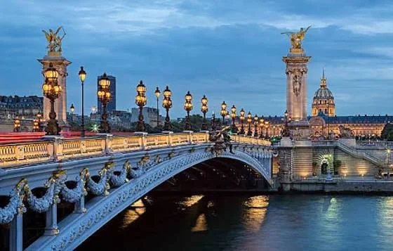 View of Alexander III Bridge over the River Seine in Paris, illuminated with streetlights and decorative sculptures, with the Invalides dome in the background at dusk.