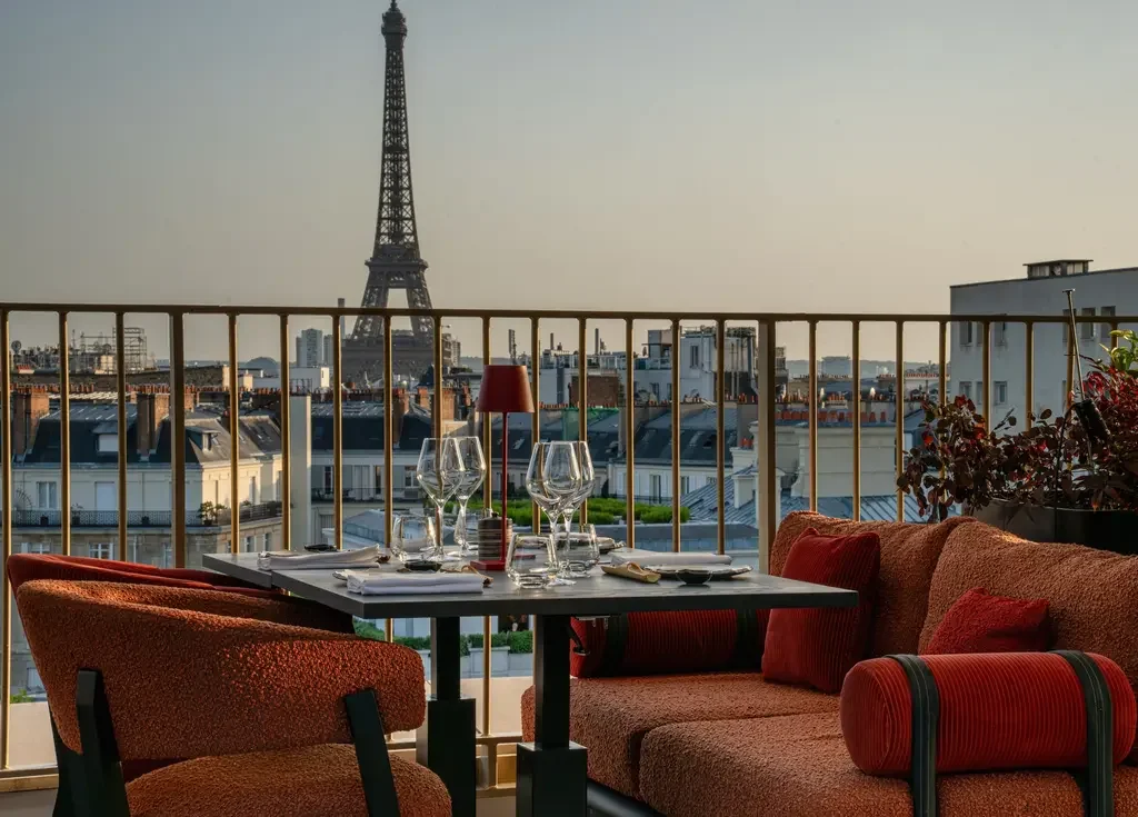 A rooftop dining area overlooking Paris with the Eiffel Tower in the background, featuring a table set with glasses and cutlery, surrounded by cozy red upholstered seating andpotted plants.