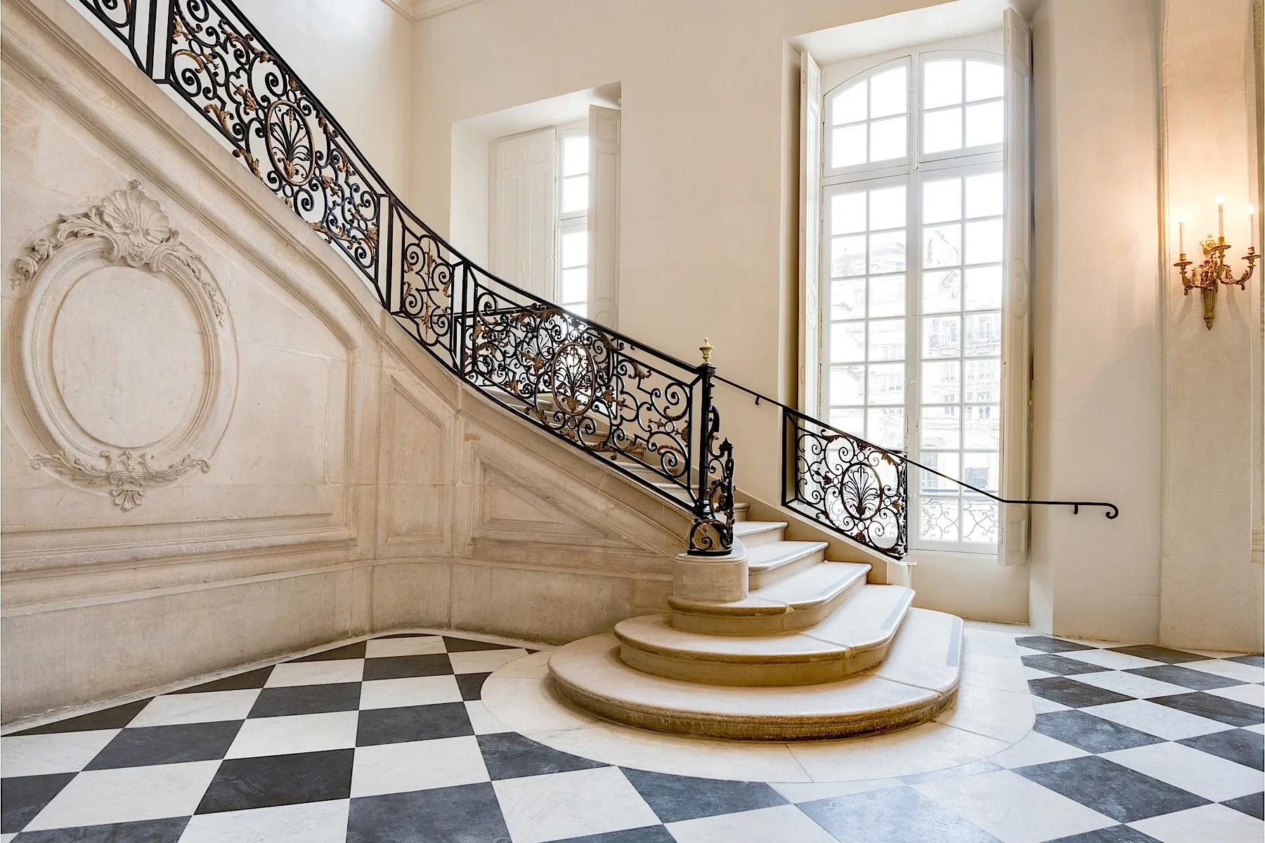 Elegant marble staircase with a decorative wrought iron railing, large windows, and checkered black and white flooring in a bright, classic interior space.