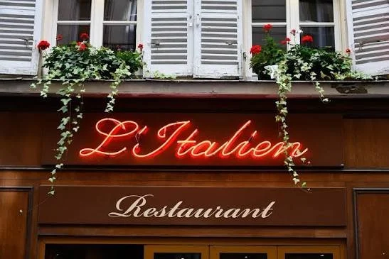 Neon sign reads 'L'italien' above the word 'Restaurant,' with flower boxes containing red and white flowers hanging from a balcony above.