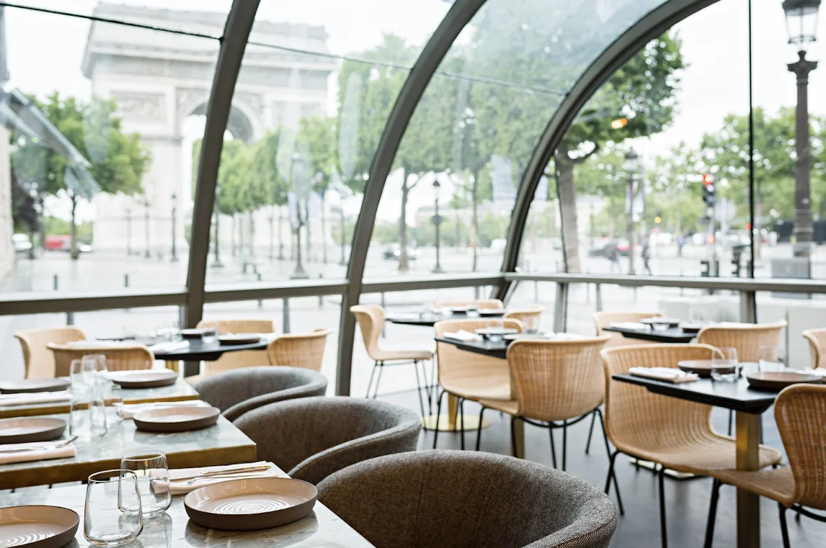 Empty restaurant with tables set for dining, large window view of a city street with trees and the Arc de Triomphe in the background.