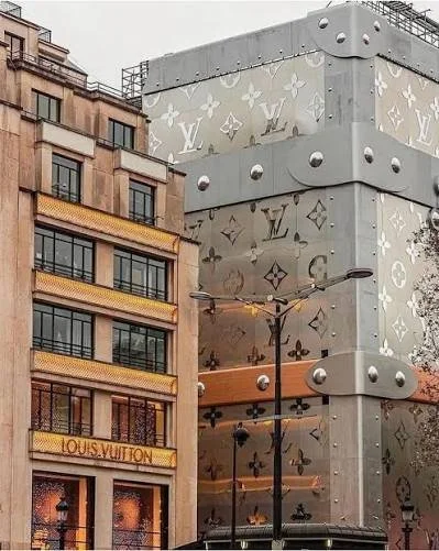 Multi-story building with a Louis Vuitton store on the ground floor, featuring large glass windows and a sign, neighboring a modern structure with Louis Vuitton monogram patterns on gray panels and rounded metal accents.