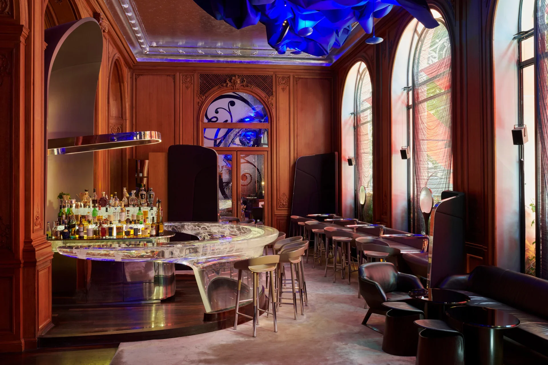 A bar with a marble countertop, bottles of alcohol, and modern black chairs inside a room with wooden paneling, large arched windows with curtains, and artistic ceiling decor with blue lighting.