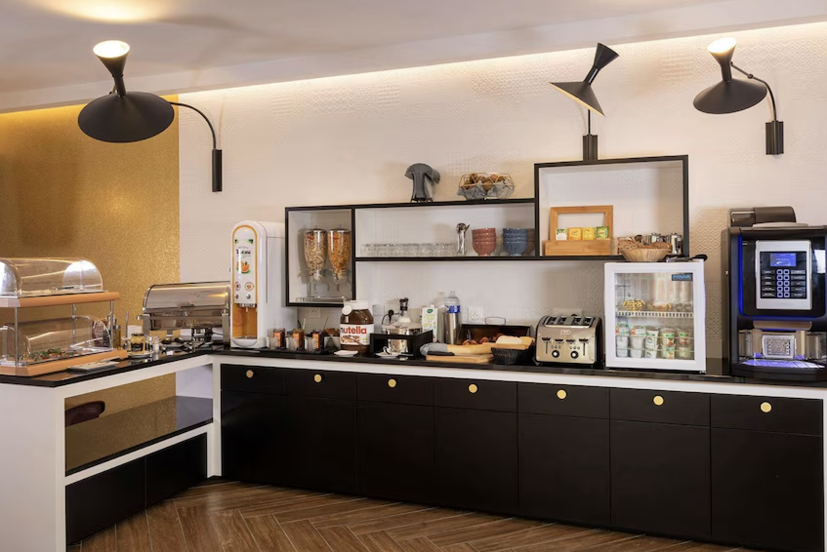 Breakfast buffet area with cereal dispenser, Nutella jar, toaster, mini fridge, coffee machine, and food warmers on a black and white counter with black drawers and gold knobs, black wall-mounted lamps, and a textured white and gold wall in the background.