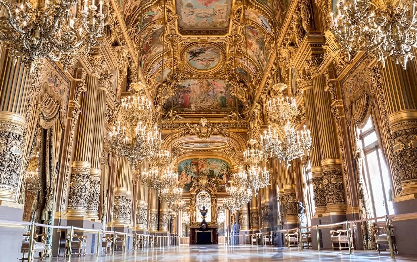 Opulent gold-embellished interior of a palace or castle with ornate gold chandeliers, detailed moldings, large windows, and ceiling frescoes.