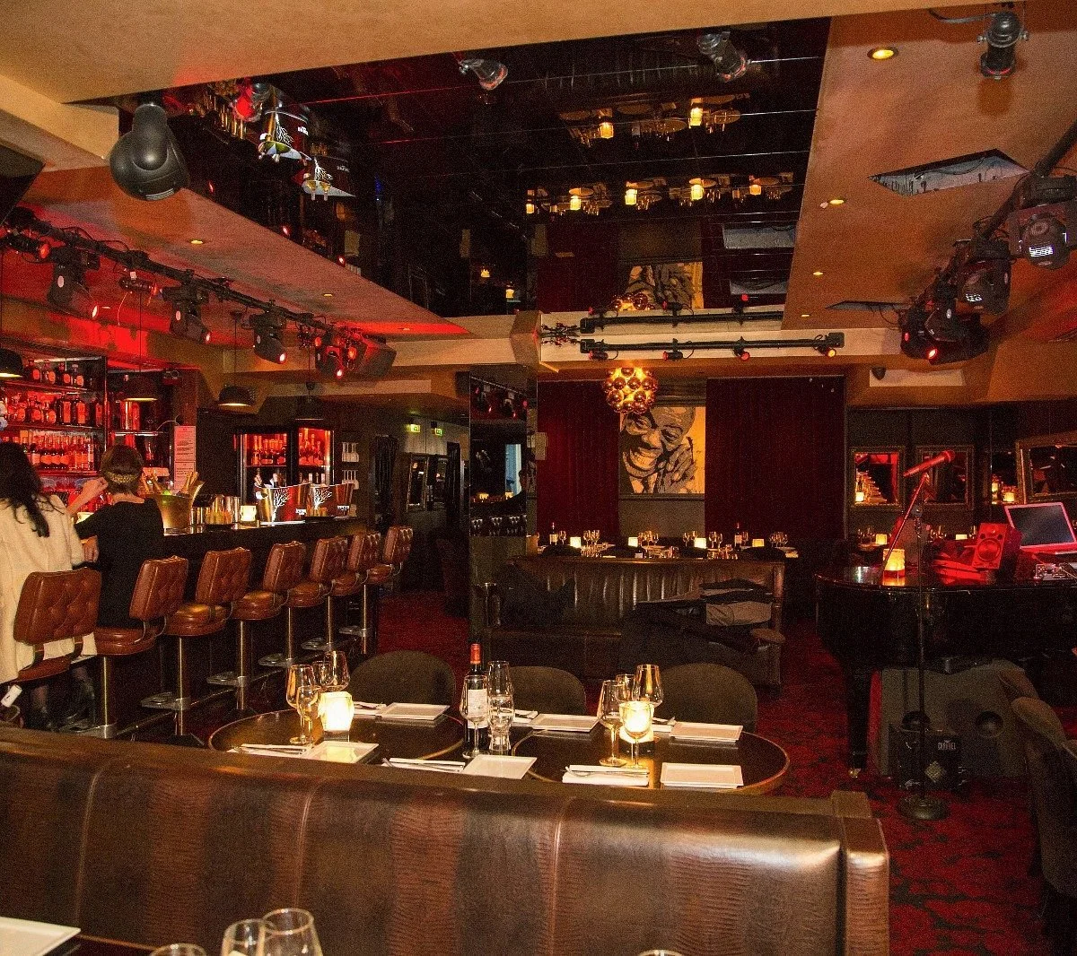 Interior of a dimly lit upscale bar and restaurant with a music stage, tables set with candles and glasses, a piano, and a bar with barstools, all decorated in warm tones and dark furniture.