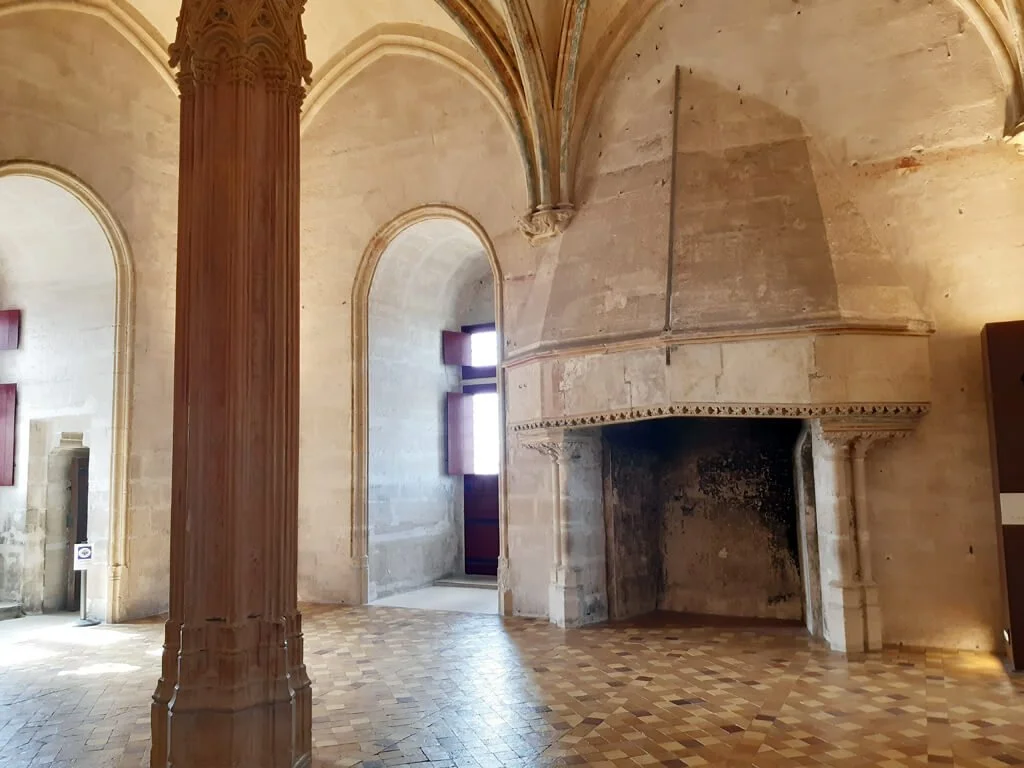 Interior of a historical building with stone walls, a large fireplace, a tall wooden column, arched windows, and a vaulted ceiling.