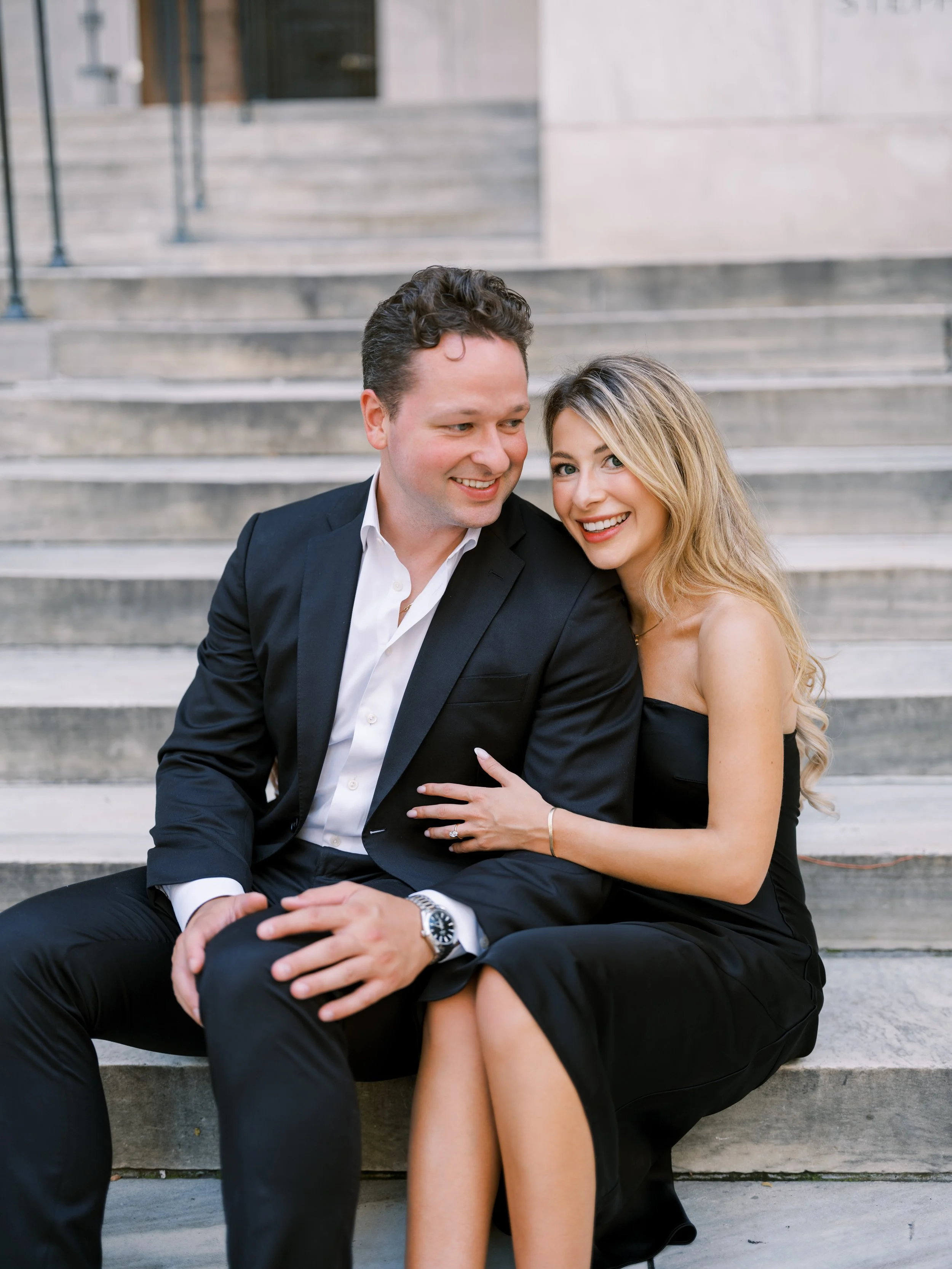 A happy man and woman sitting on concrete steps outside. The man is in a black suit and white shirt, and the woman is in a black strapless dress. They are smiling and the woman has her hand on the man's knee, showing her engagement ring.