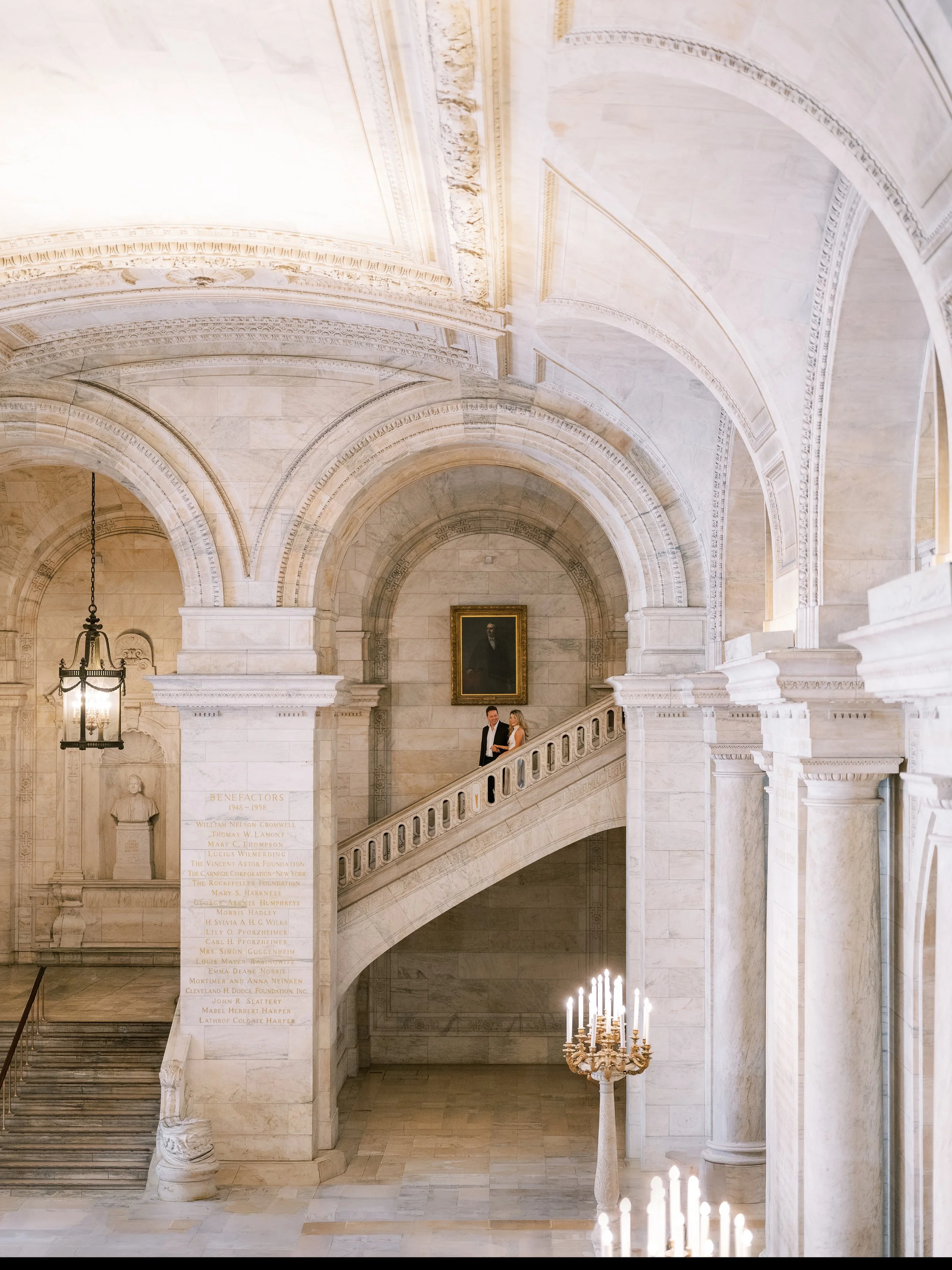 Interior of a grand marble building with arched ceilings, a staircase, and a chandelier, featuring a portrait on the wall and a list of names on the pillar. New York Public Library. NYPL. Samantha Daniell & Michael Moore. 