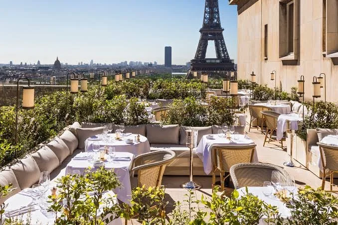 Outdoor dining area on a rooftop in Paris with tables set for a meal, surrounded by greenery, with the Eiffel Tower and Paris skyline in the background.