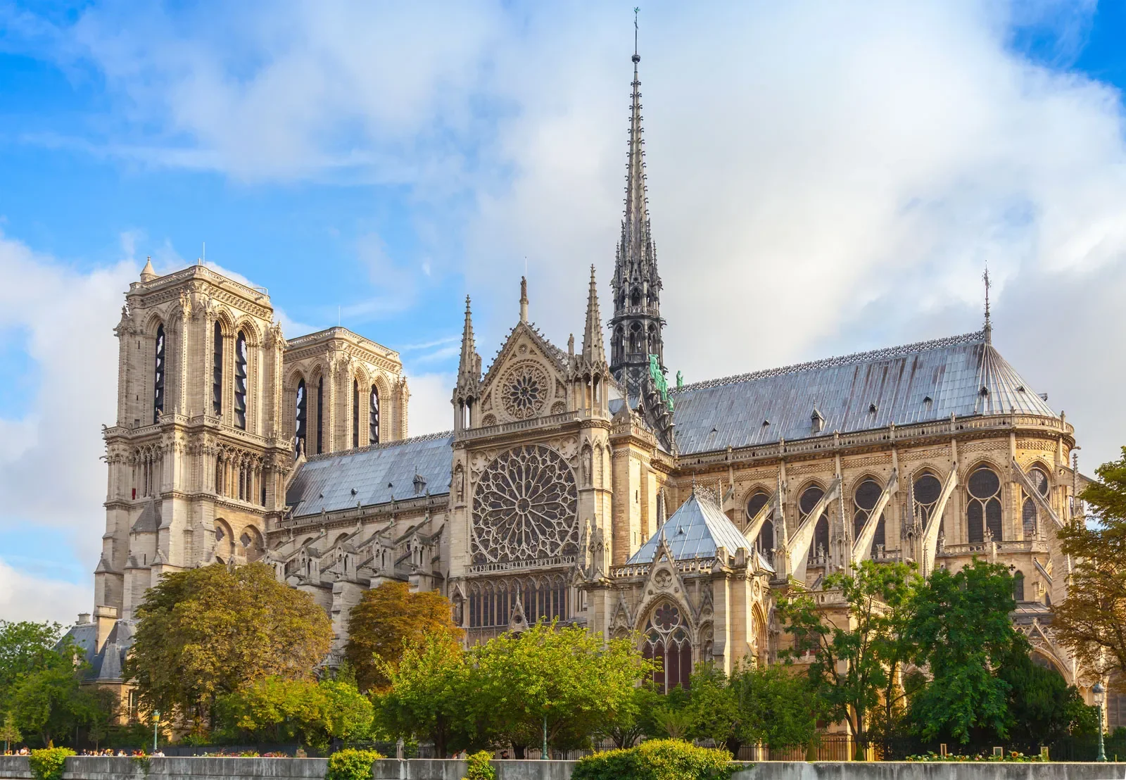 The Notre-Dame Cathedral in Paris, France, shown from the front, featuring its Gothic architecture, twin towers, large rose window, and spire, with trees in the foreground and a partly cloudy sky.