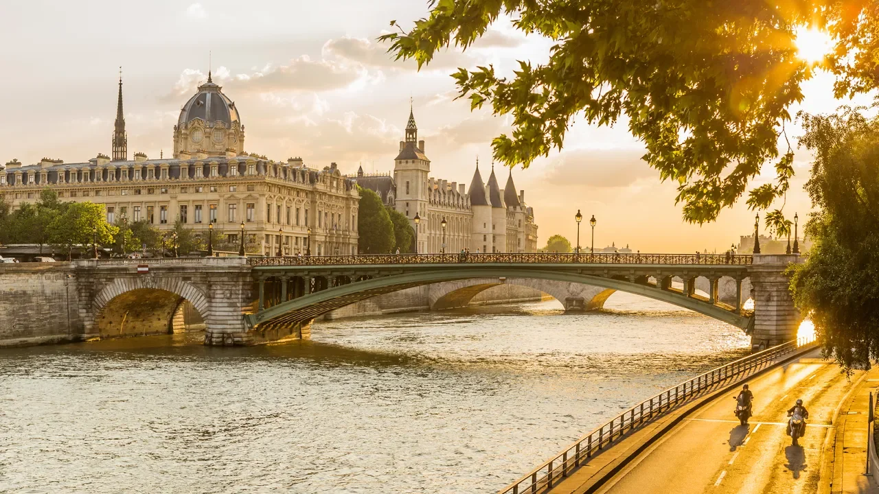 View of a historic bridge over the river with classic Parisian architecture and a cloudy sky in the background, during sunset, with two motorcyclists riding along the riverbank.