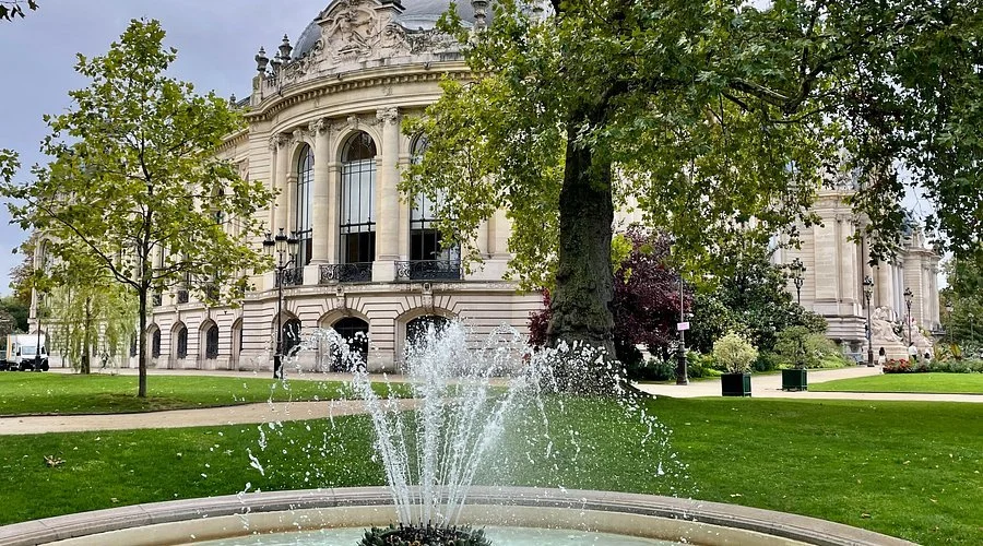 A fountain in a park in front of a historic building with large windows and ornate architecture, surrounded by green trees and grass.