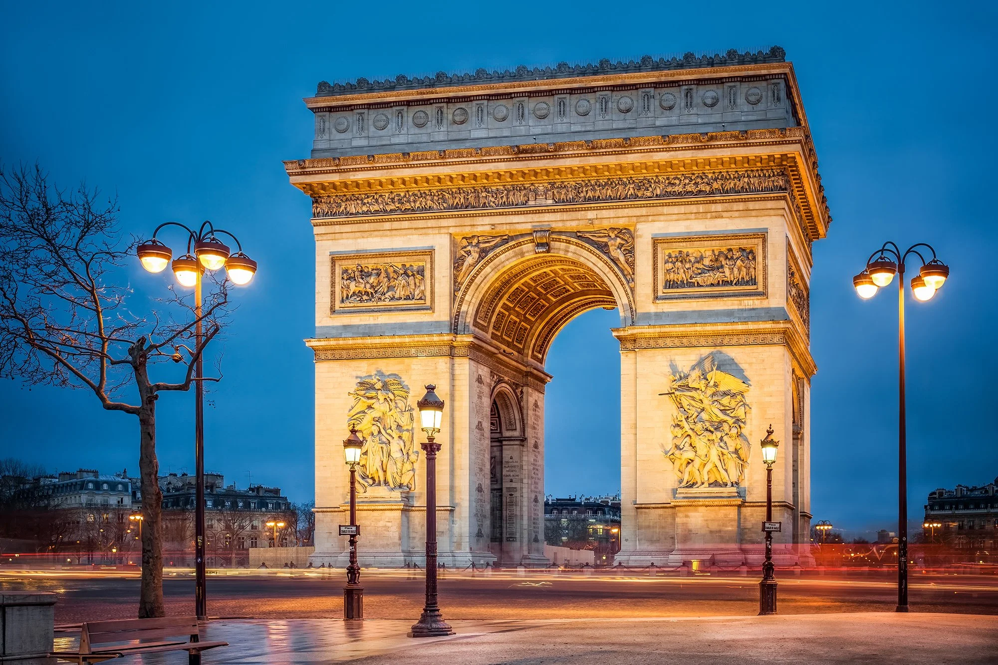 The Arc de Triomphe in Paris illuminated at dusk, with streetlights and bare trees in the foreground.
