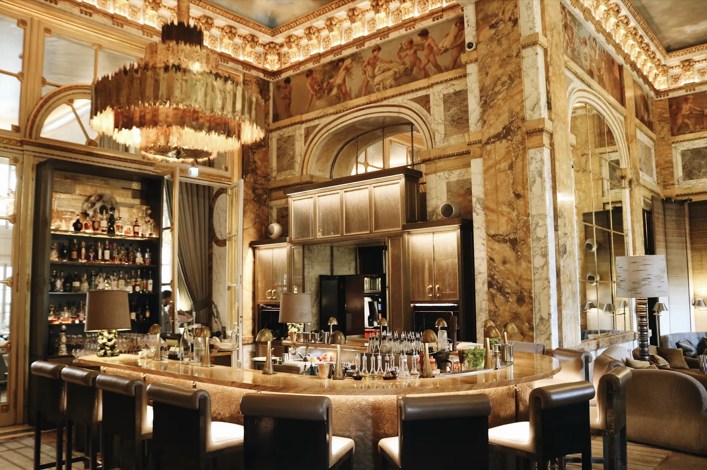 Elegant interior dining area with marble walls, gold accents, and a large chandelier, featuring a bar with bottles and tables with chairs.