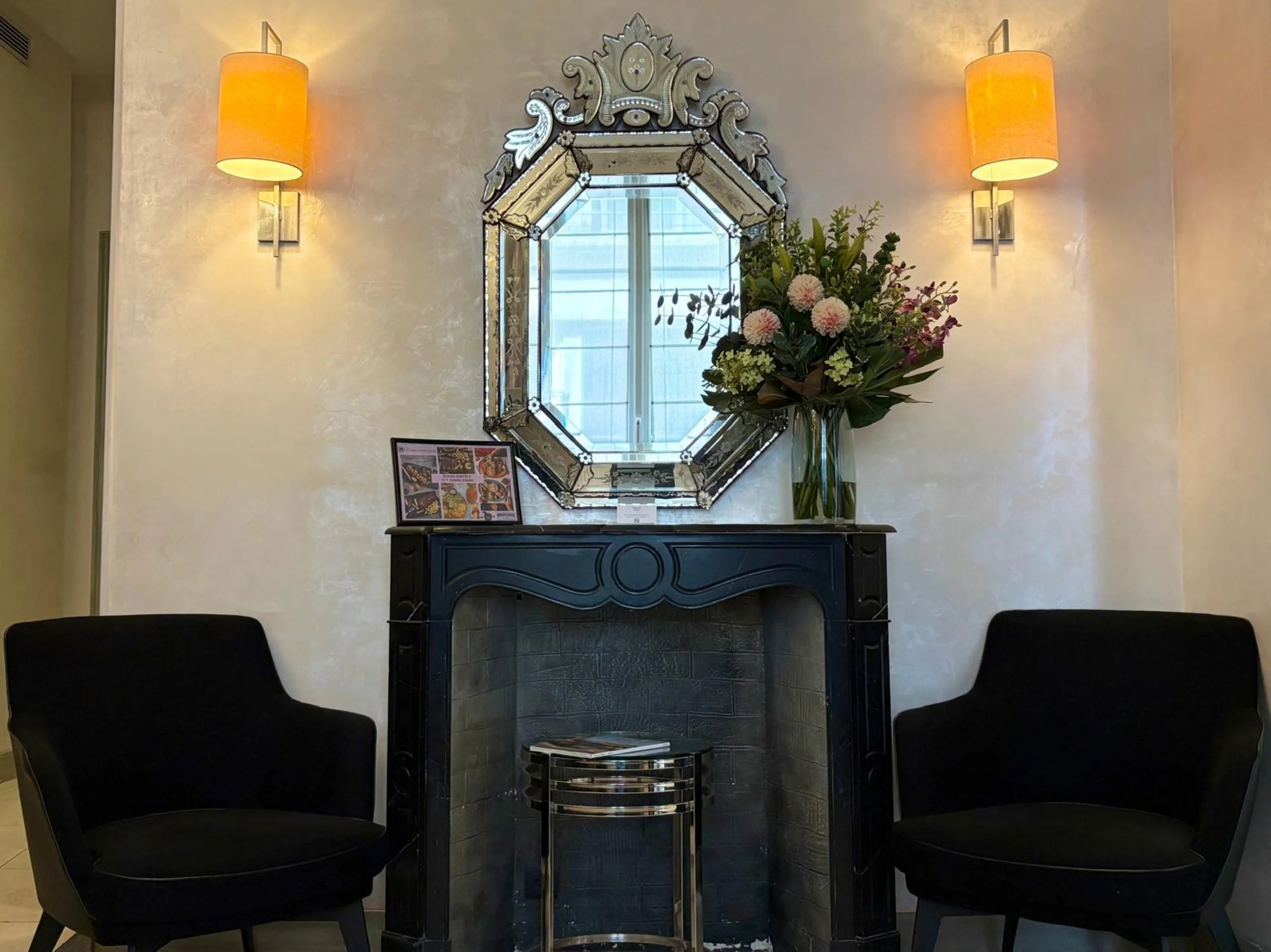 A decorative wall mirror with a floral arrangement in a glass vase placed on a dark-colored table below the mirror. Two wall-mounted lamps with yellow shades illuminate the beige textured wall.