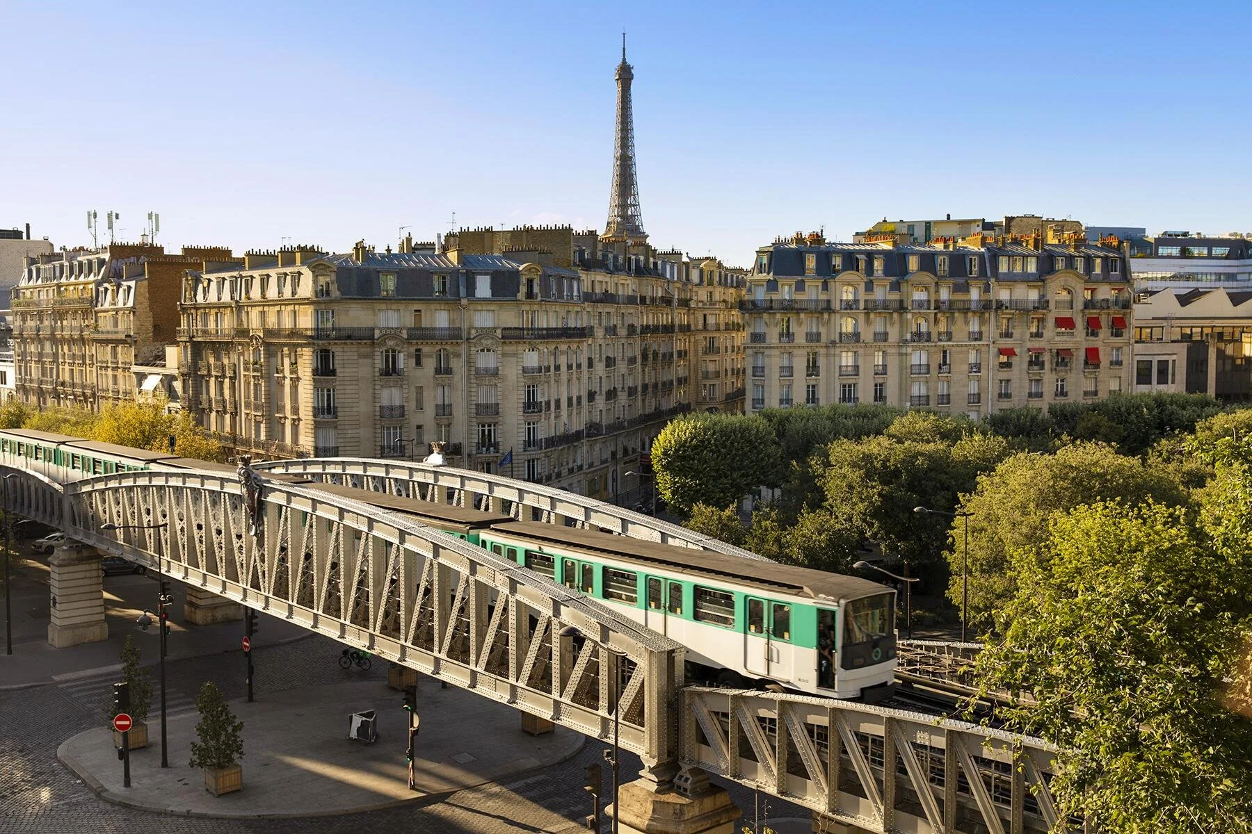 Paris cityscape with Eiffel Tower in the background, classic Parisian buildings, a green and white metro train on an elevated track, with trees and street below on a sunny day.