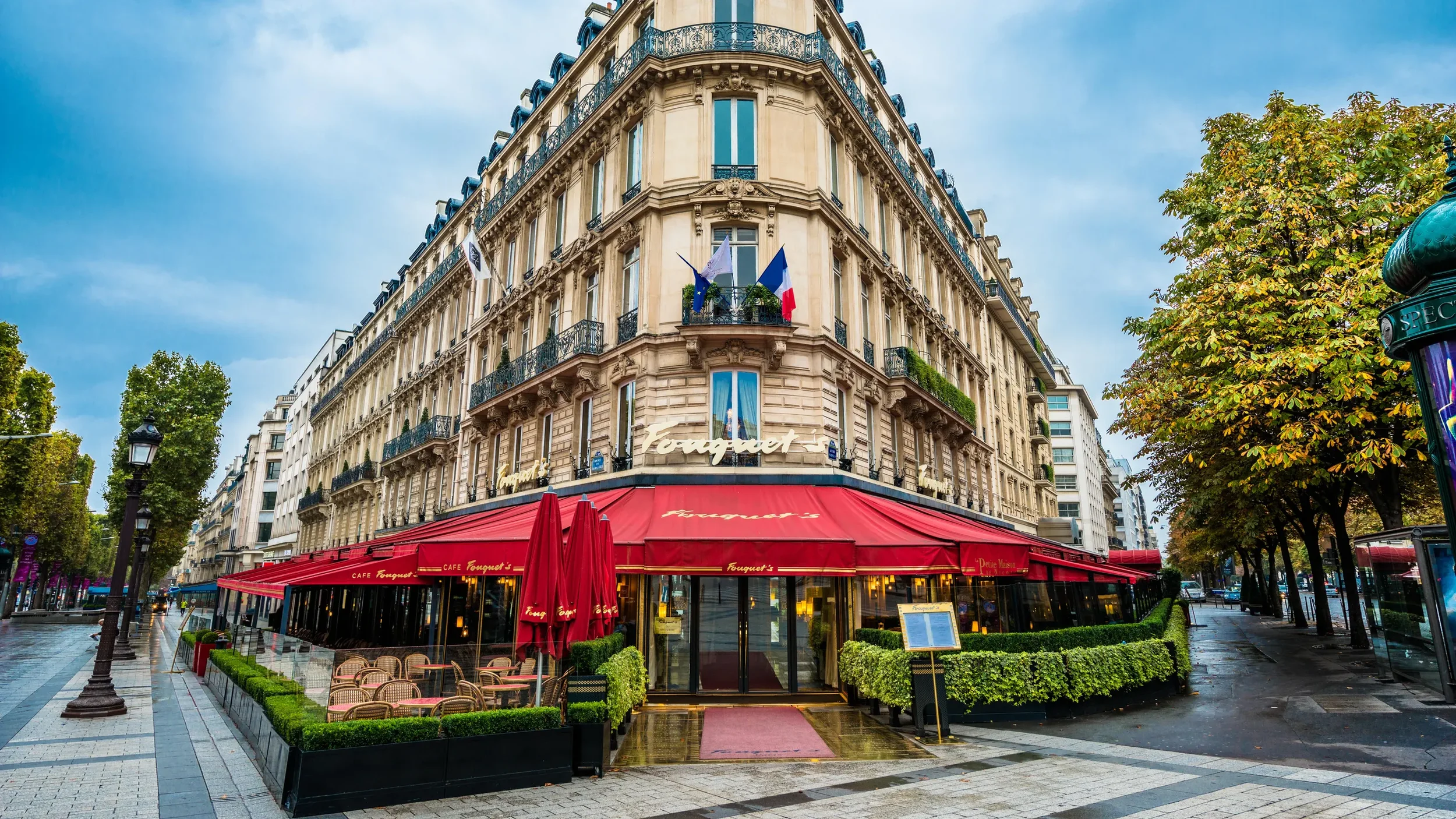 A Parisian cafe named Fouquet's on a corner building with ornate architecture and flags, outdoor seating area with red umbrellas, and trees lining the street on a rainy day.