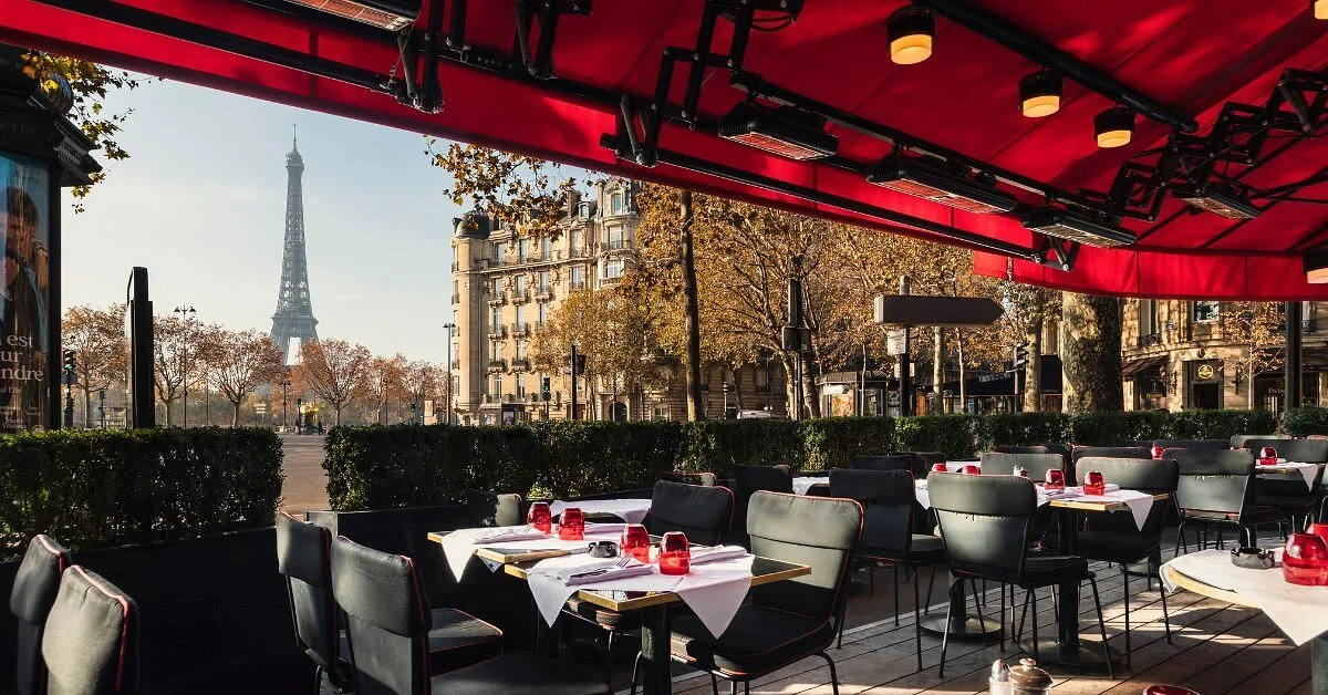 Empty outdoor Parisian restaurant with tables set with white tablecloths, red glasses, and black chairs, overlooking Paris cityscape with Eiffel Tower in the background on a sunny day.
