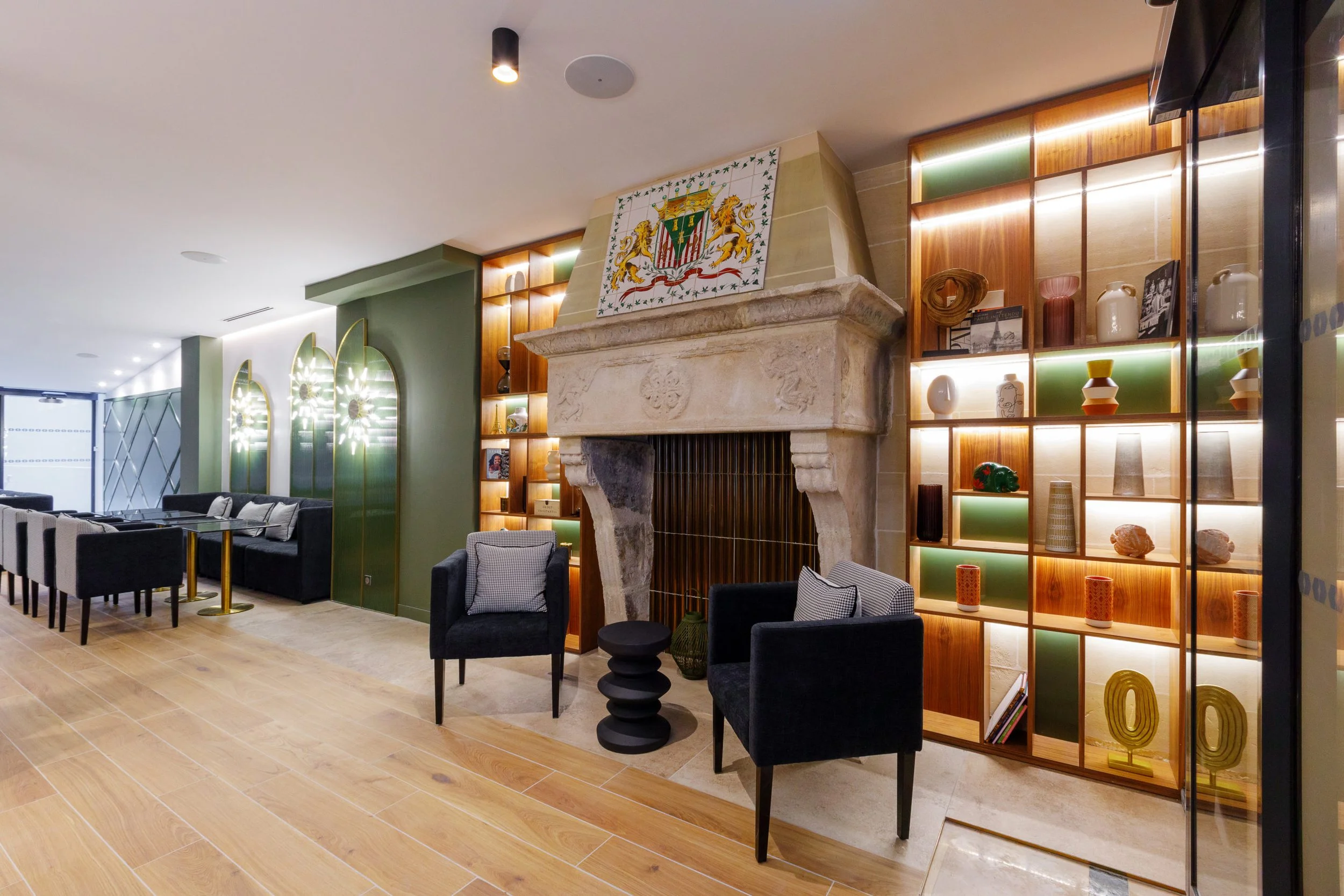 Interior of a modern living room with a fireplace, a green wall with decorative mirrors, and a wooden bookshelf with various vases and books, illuminated by soft lighting.