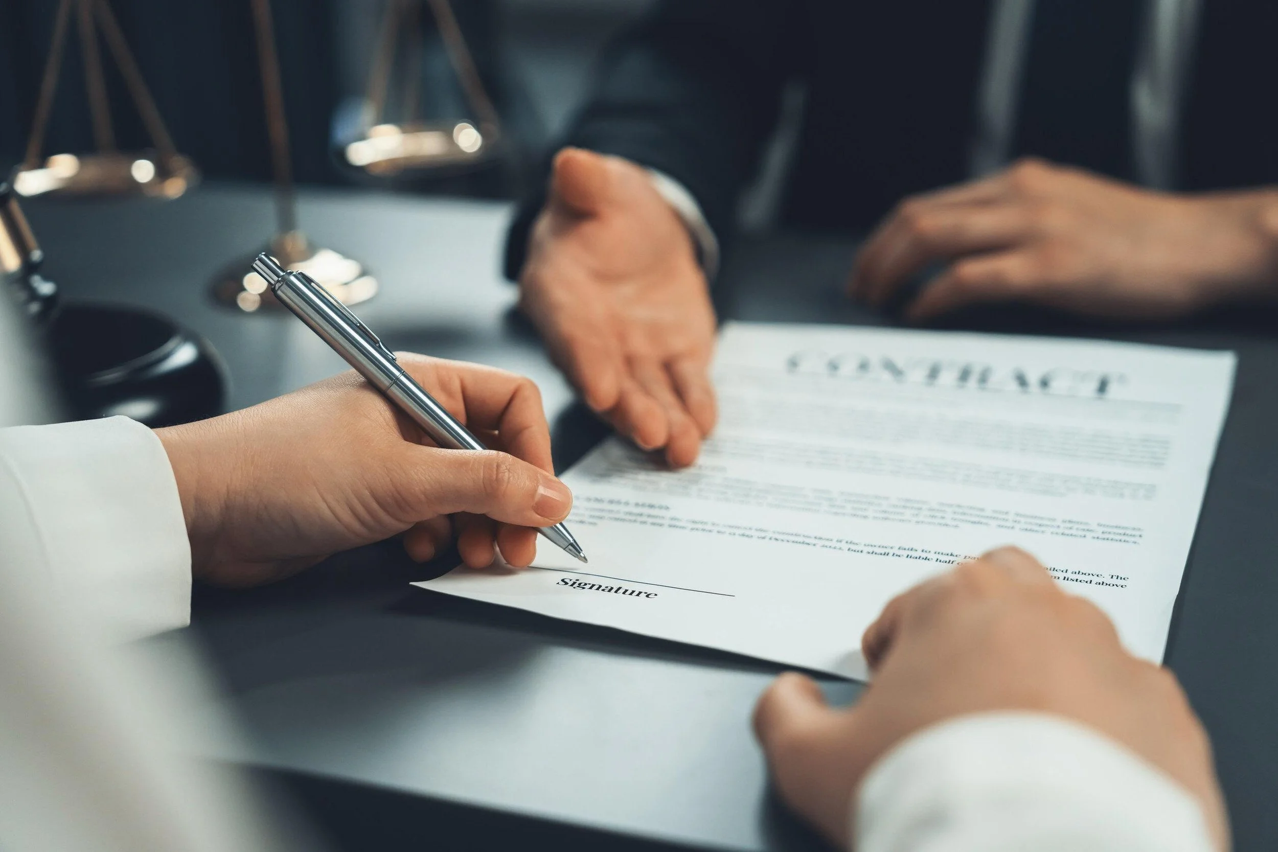 A person in a business suit signs a document with a silver pen, with the word 'signature' at the bottom of the paper. Another person, also in business attire, is seated at the table with hands visible, and a pair of scales can be seen in the background.