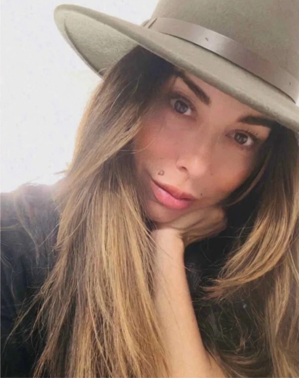 A woman with long, wavy hair wearing a beige wide-brimmed hat and a black top, posed with her hand resting under her chin, looking into the camera.