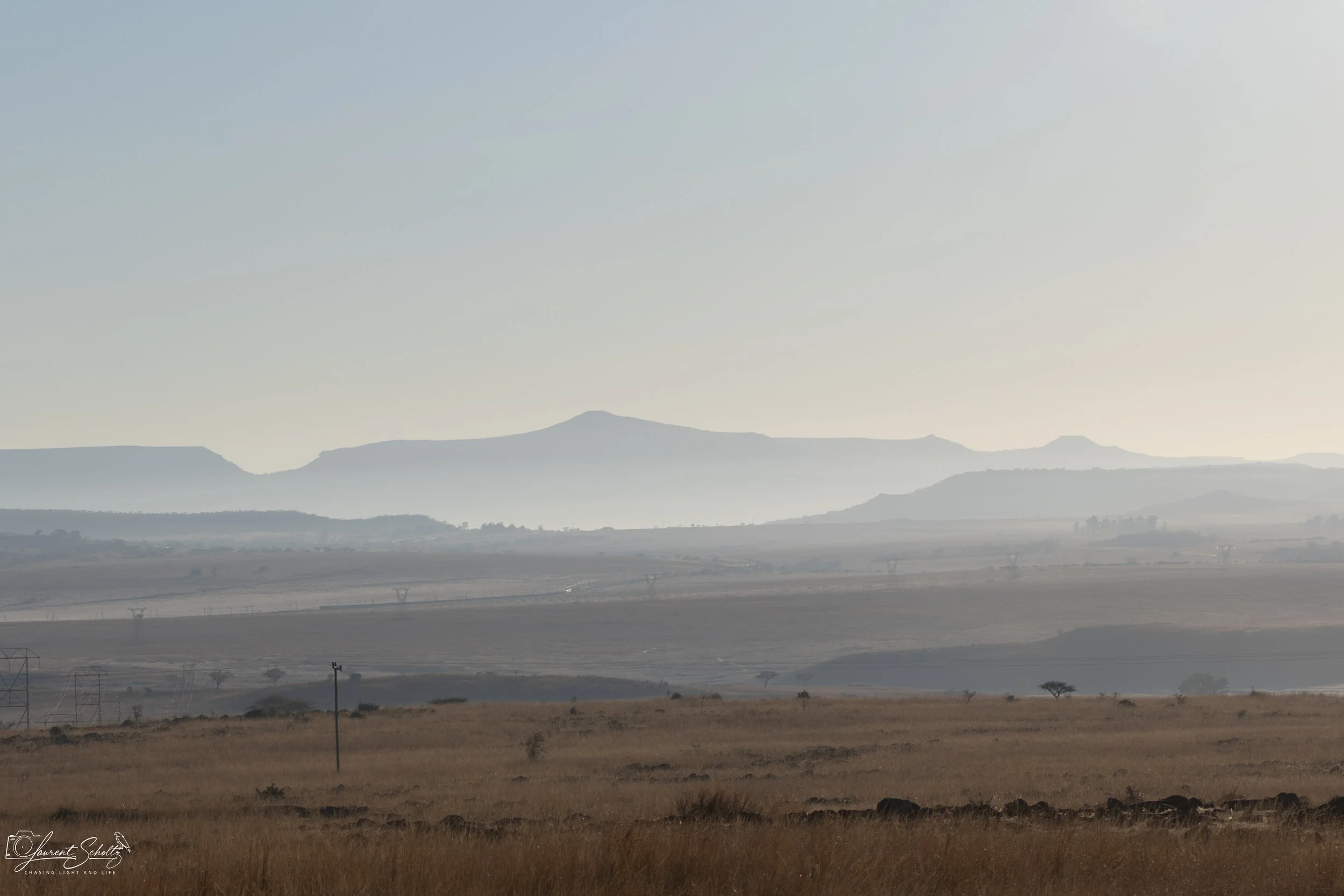 A landscape view of distant mountains with a layer of haze, rolling hills in the foreground, a few scattered trees, and power lines.