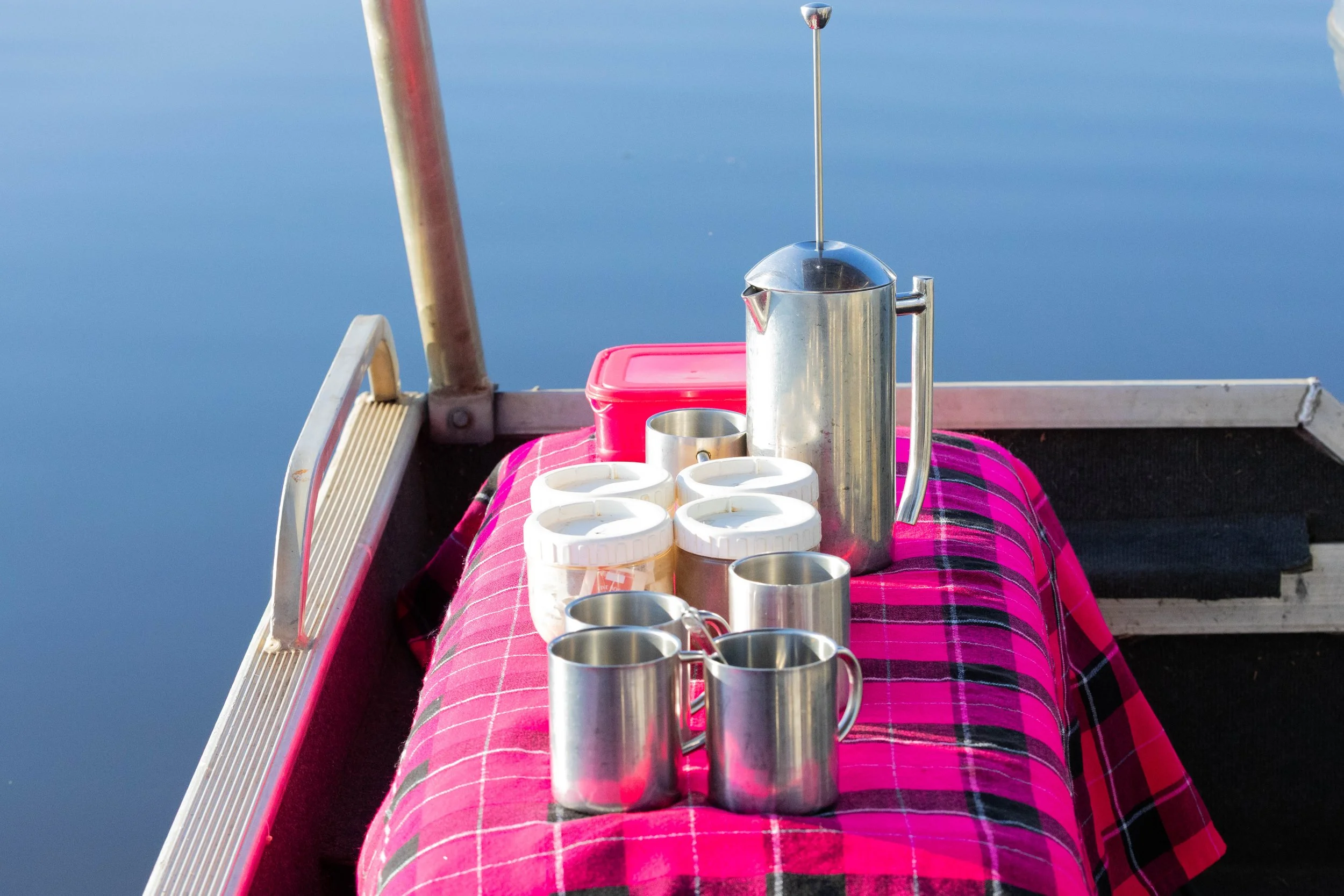 A table with coffee mugs and a thermos on a pink checkered cloth, set on a boat with blue water in the background.