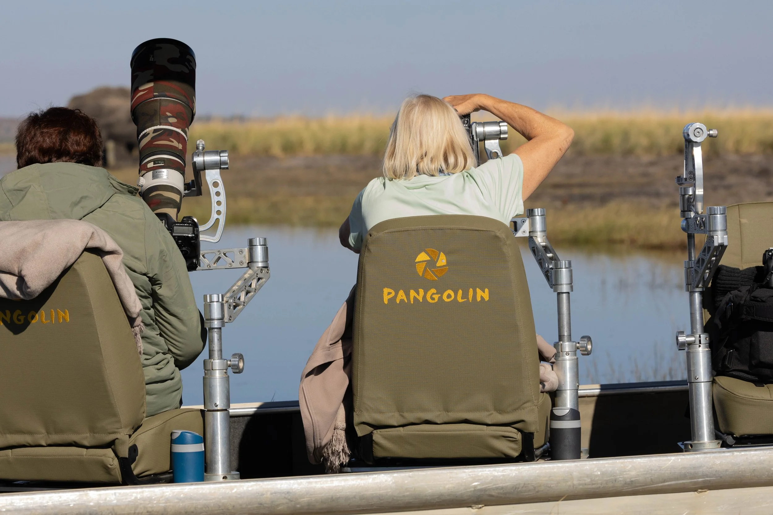 Two women and a man seated on a boat with metal railings, using telescopes to observe a distant landscape with grassy marshes and a large rock formation, under a clear sky. The woman in the middle has the name "Pangolin" embroidered on her green jack