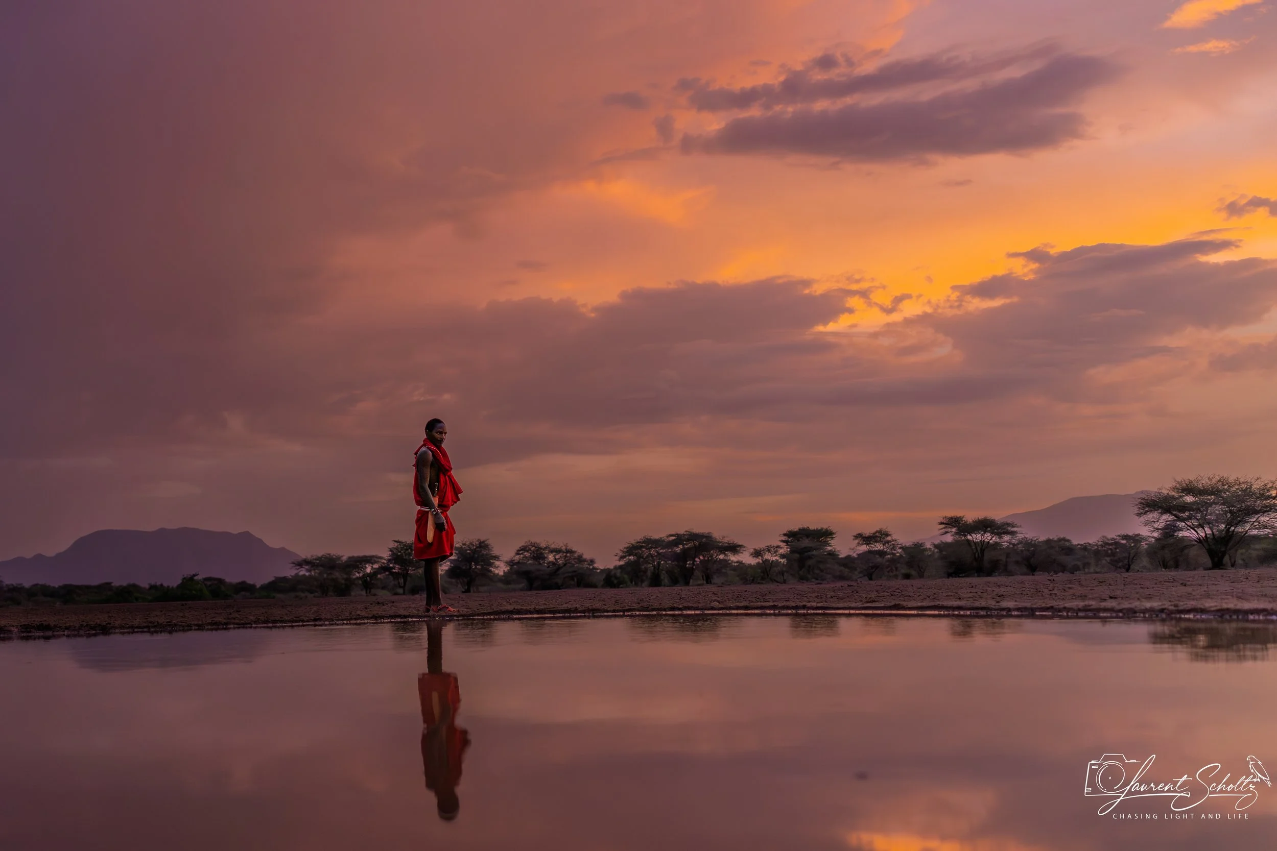 A solitary person wearing a red garment standing by a reflective body of water at sunset, with a mountain range and scattered trees in the background.