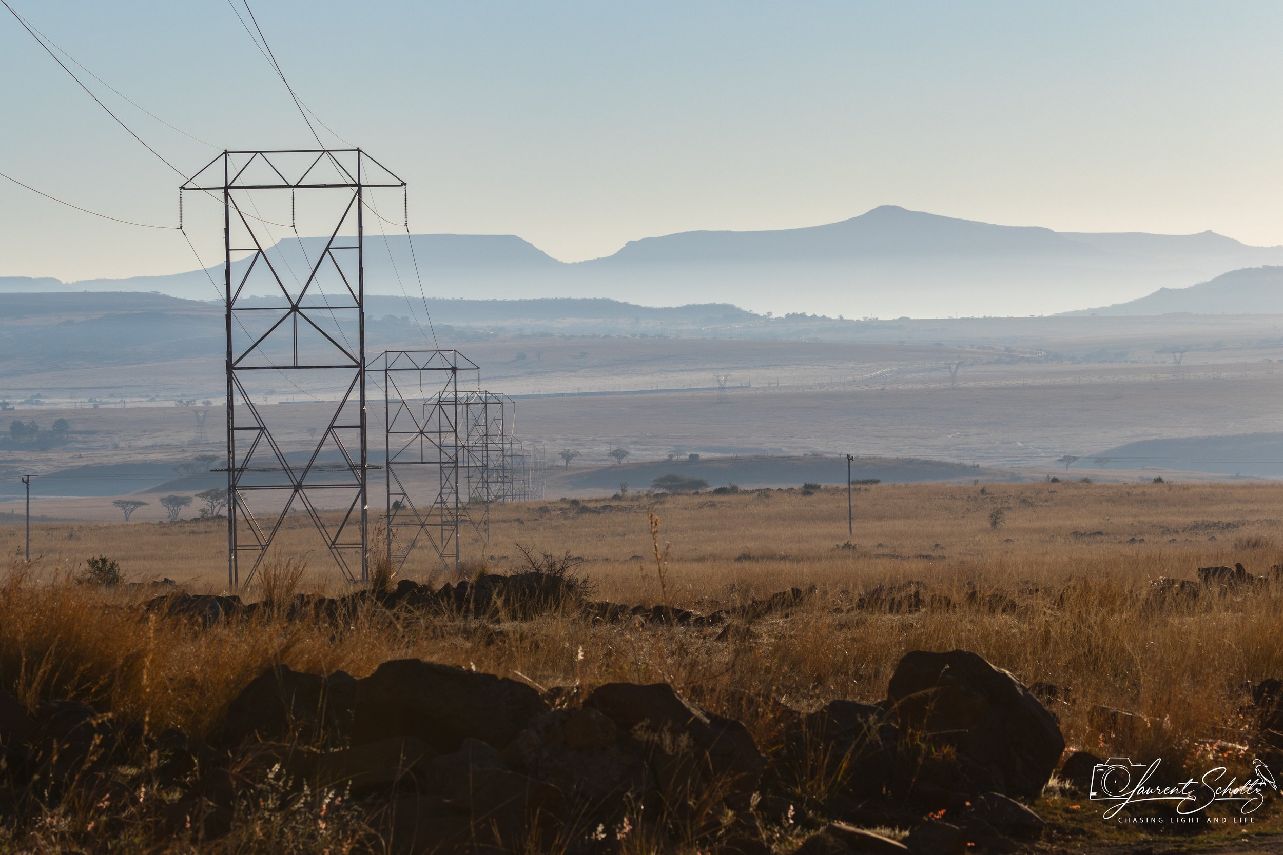 A landscape scene featuring power lines and towers stretching across a vast, open field with rocks and tall grasses in the foreground. In the background, there are rolling hills and distant mountains under a pale sky.
