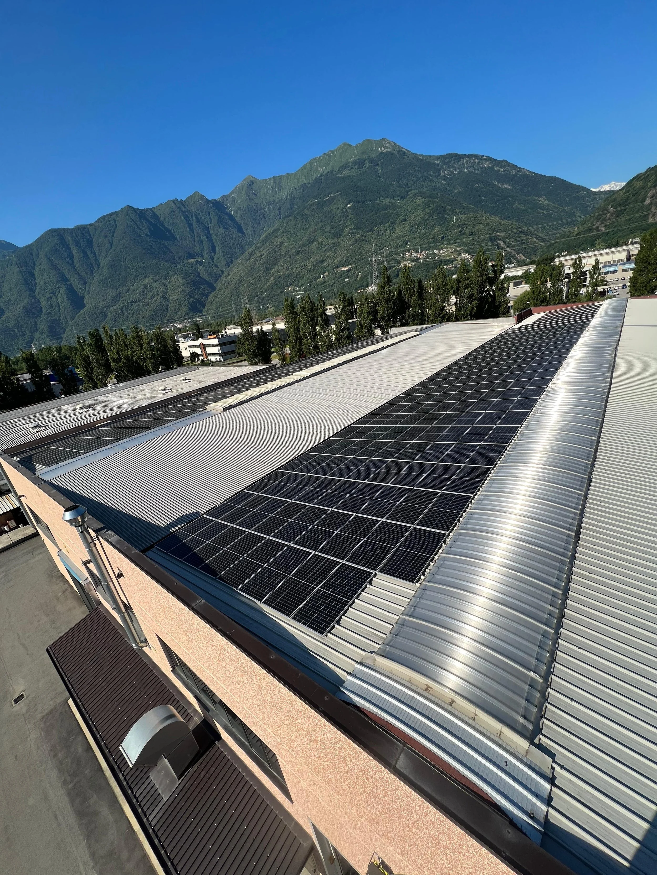Tetto di un edificio con pannelli solari e montagne verdi sullo sfondo, cielo chiaro e sole.