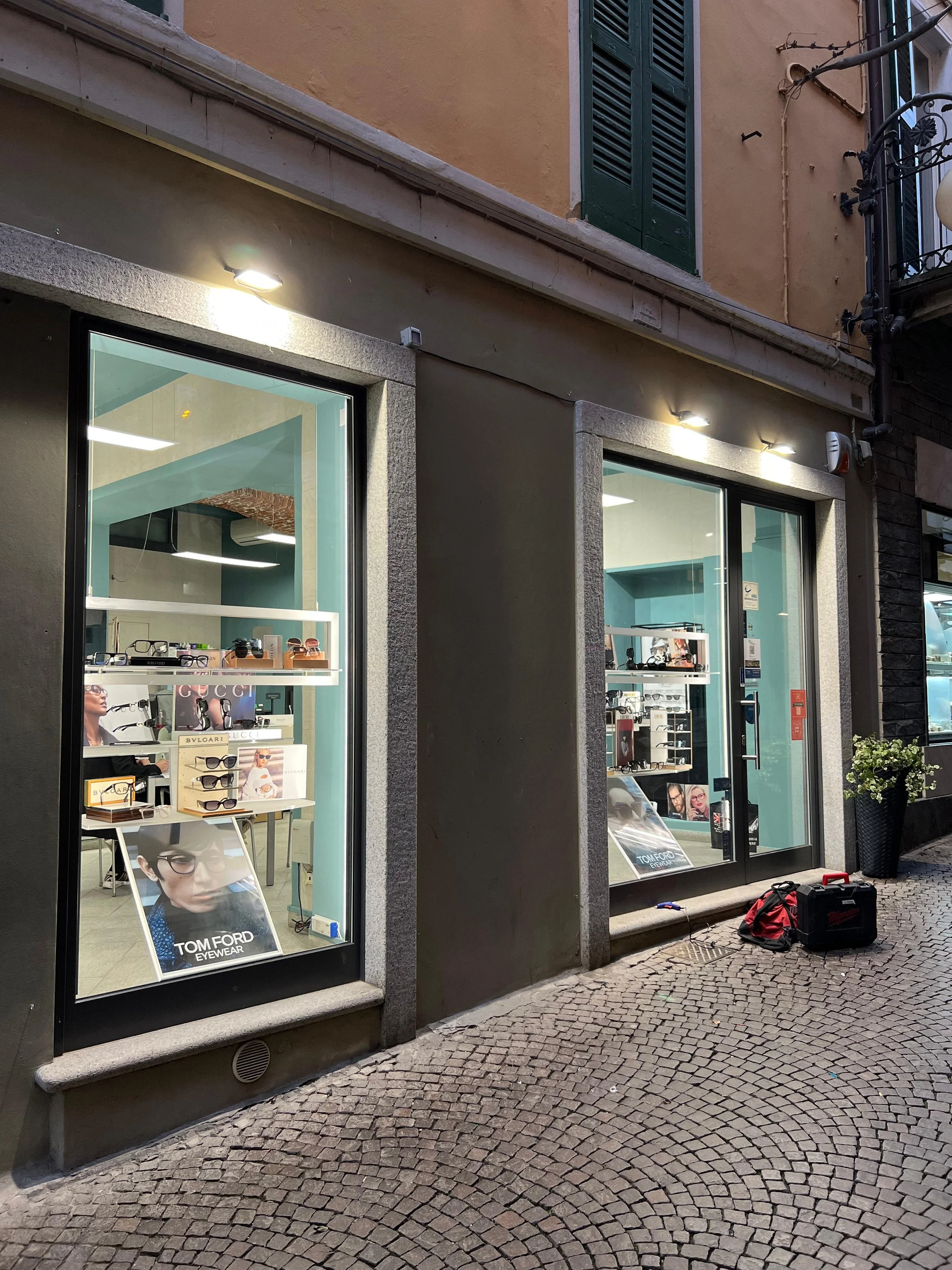 Vetrina di negozio di occhiali con poster pubblicitario di Tom Ford, ambientato in strada con pavimentazione in cobblestone, presenza di lampade esterne e piante.