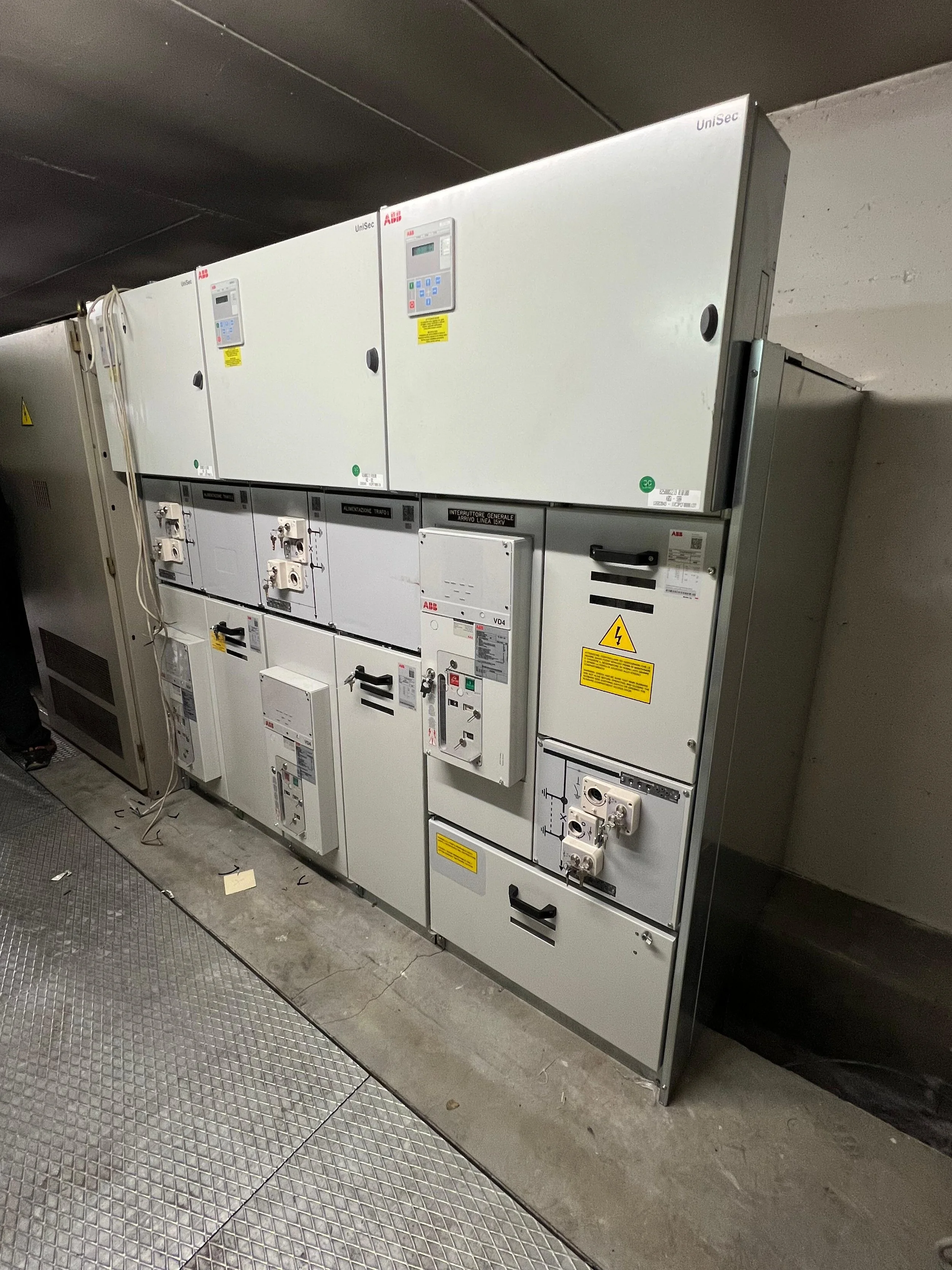Armadio di apparecchiature di controllo e distribuzione elettrica, di colore grigio, con varie porte, interruttori e indicazioni, posizionato su un pavimento in piastrelle metalliche in ambiente industriale o tecnico.