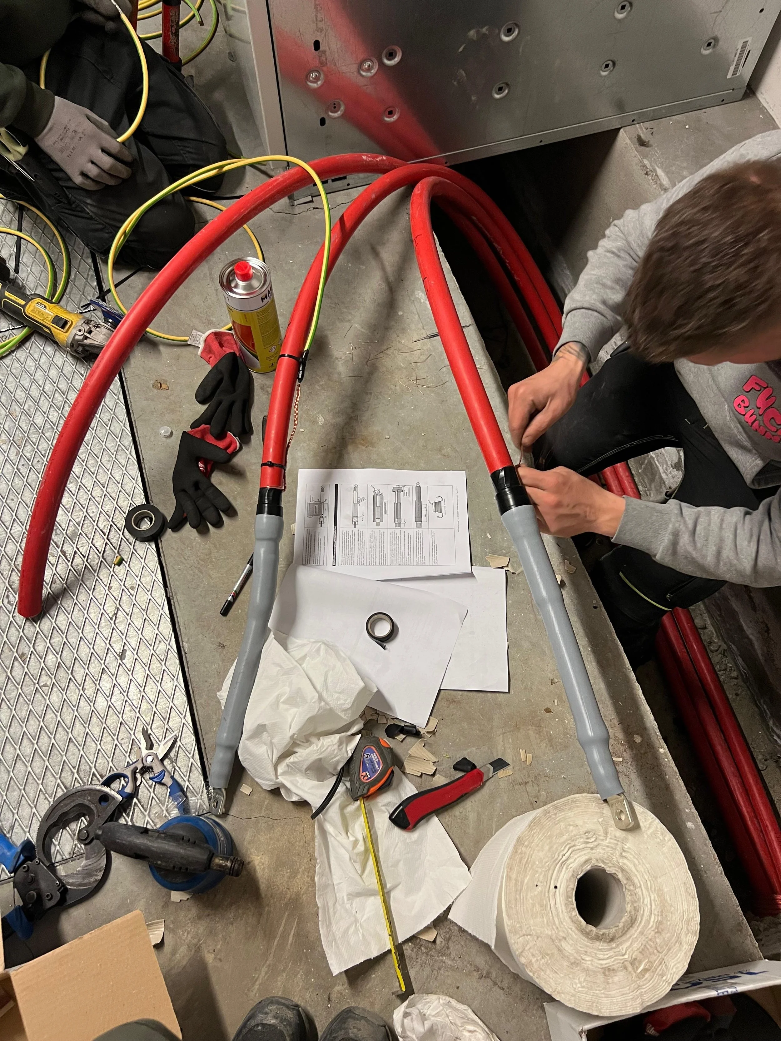 Immagine di un'officina con due persone che lavorano su un telaio di bicicletta o veicolo, con vari strumenti, cavi rossi e gialli, un tubo di schiuma e una serie di parti meccaniche sparse sul pavimento.