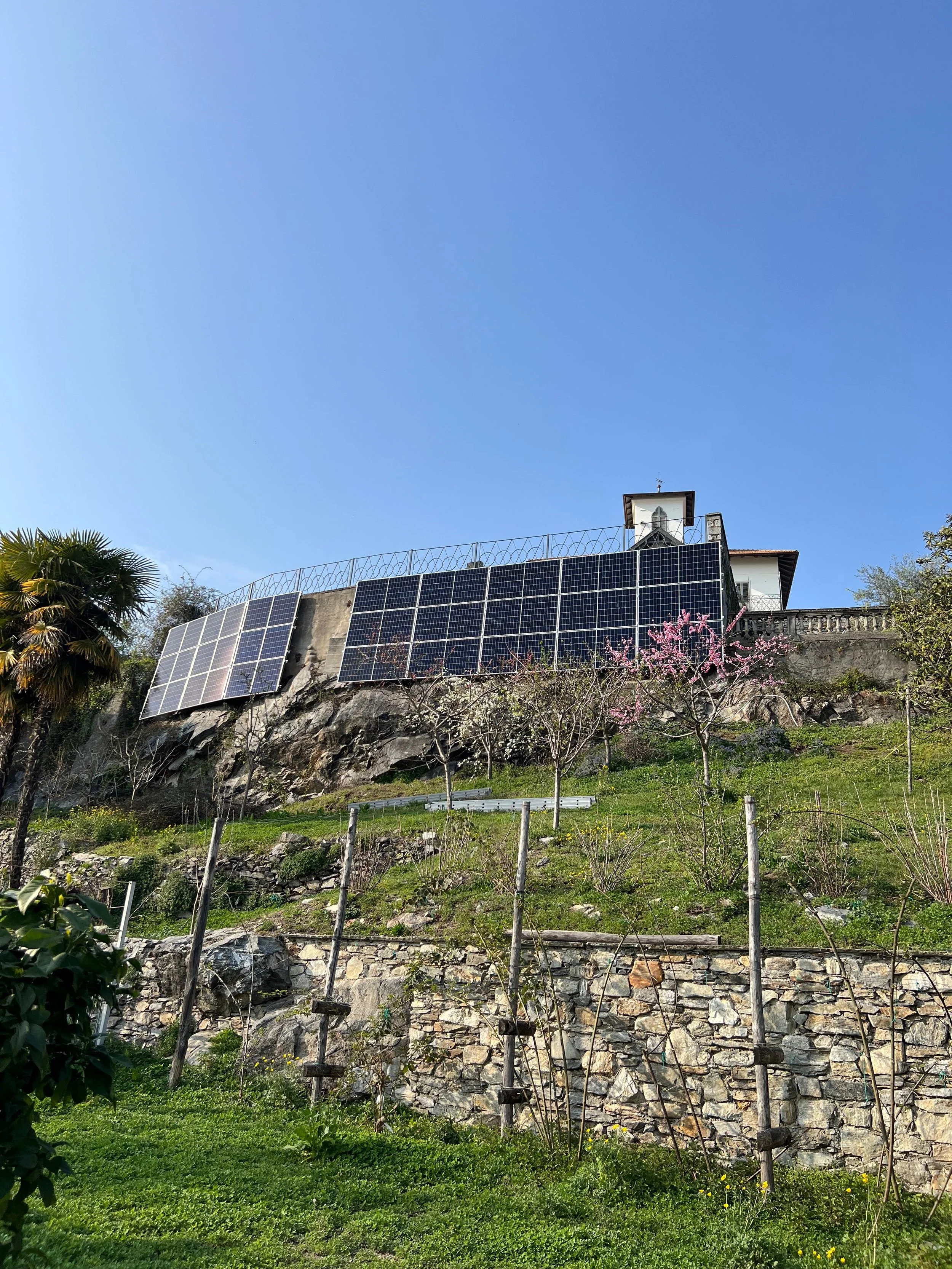 Casa su una collina con pannelli solari sul tetto, alberi e fiori in primo piano, sotto un cielo chiaro e blu.