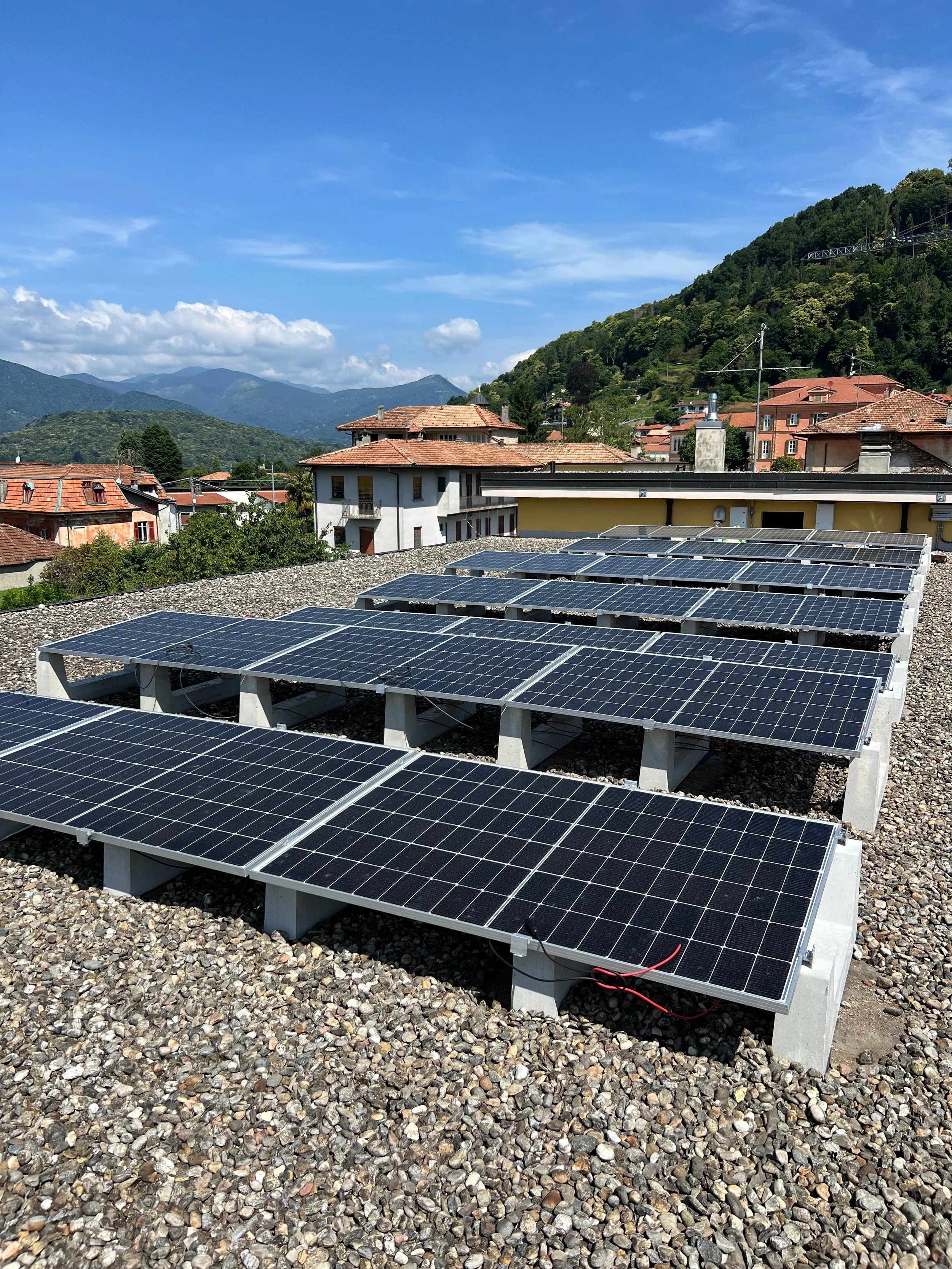 Pannelli solari installati su un tetto di un edificio con una città sullo sfondo e montagne nella distanza, sotto un cielo sereno con alcune nuvole.
