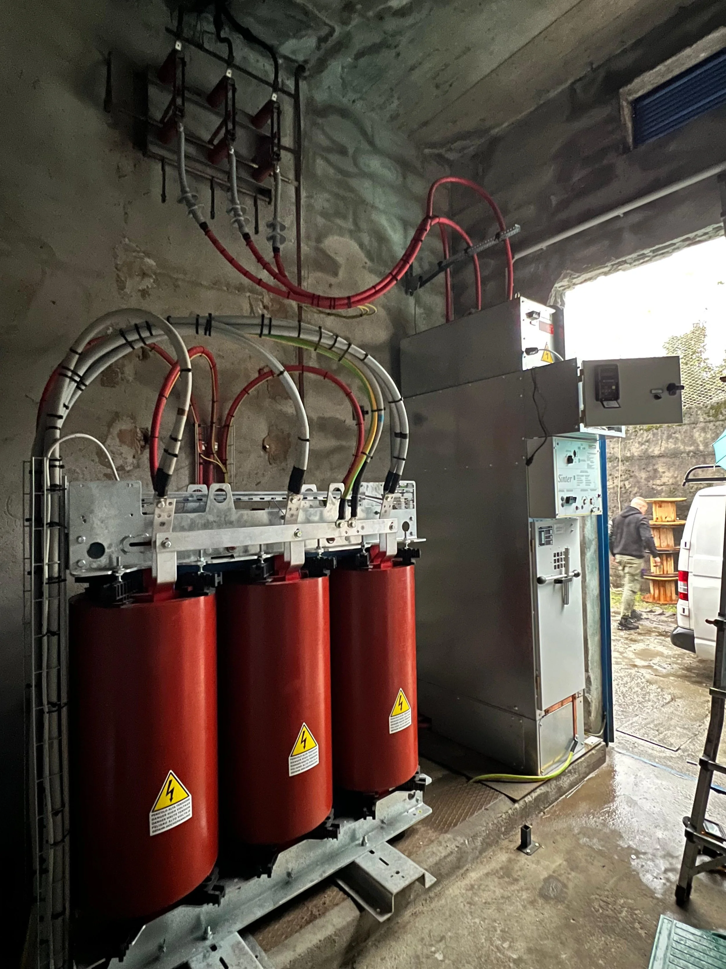 Immagine di un impianto di trasformatori elettrici installato in un locale, con tubi e cablaggi, pareti di cemento e accessori di sicurezza.